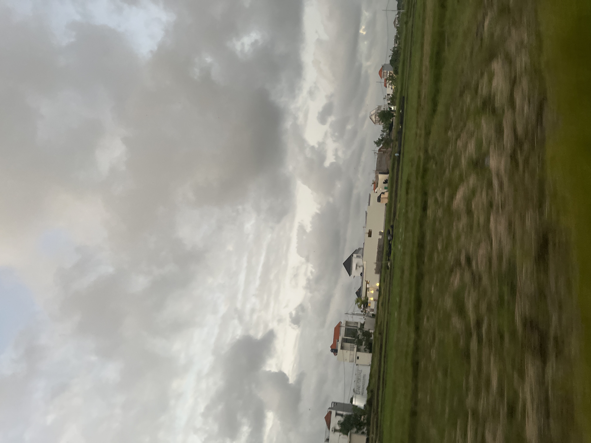 The image depicts a village in Mengwi, Indonesia, under a cloudy sky, possibly before a storm. The scene is oriented vertically, suggesting it was captured from a moving vehicle or tilted camera.
On the left, the sky is filled with dramatic, heavy grey clouds, with patches of lighter sky peeking through. Scattered throughout the sky are small, dark specks, which could be birds or insects.
To the right of the sky, a line of modern houses is visible. These houses are mostly white with some featuring orange or red roofs. Some have multiple stories and balconies. One house has a prominent black, pyramidal roof. Another has a sign that appears to read "LAND FOR SALE" and possibly a phone number below it. Lights are visible inside some of the houses, suggesting it might be late afternoon or early evening.
Adjacent to the houses is a vast expanse of green grass, which appears to be a field or a large lawn. The grass is blurred due to motion, but its texture suggests it's long and possibly wild. The line between the houses and the field is distinct, with the field extending to the right edge of the frame.
The overall mood of the image is somewhat somber due to the overcast sky, but the presence of the houses and the vibrant green of the field offer a sense of life and development. The time of day is not definitively clear, but the lighting suggests it's not midday. The location is specified as Mengwi, Indonesia.