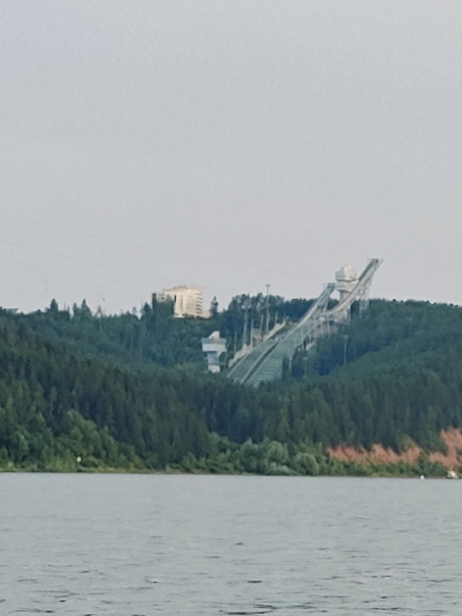 An image shows a large ski jumping complex on a wooded hill overlooking a body of water. The ski jump is a modern structure with a tall tower and a long, sloping ramp. A building, possibly a hotel or administrative building, is located on the hill behind the ski jump. The trees are predominantly coniferous, with some deciduous trees interspersed. The water in the foreground is calm, with a slight ripple. The sky is overcast and a pale gray. The scene is likely a recreational or sports facility.