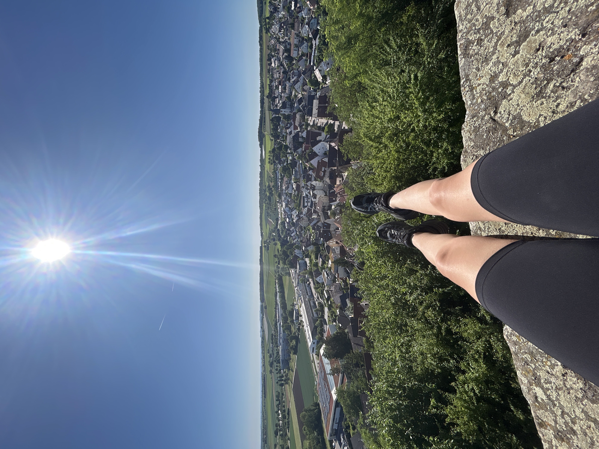 A high-angle, daytime, outdoor shot features a person's legs and feet dangling over the edge of a rocky precipice. The person is wearing black athletic shorts and black sneakers. The lower half of their legs is visible, with their feet dangling in the air. Below them is a lush green area filled with trees and vegetation. Beyond this green area is a view of the city of Untergruppenbach, Germany, characterized by densely packed buildings with varying roof colors, interspersed with trees and greenery. In the distance, the city transitions into agricultural fields. The sky above is a clear, bright blue, with a prominent sunburst in the upper left corner, casting a lens flare across the image. A faint contrail is visible in the sky near the sun. The rock surface appears to be light-colored with lichen or moss growth.