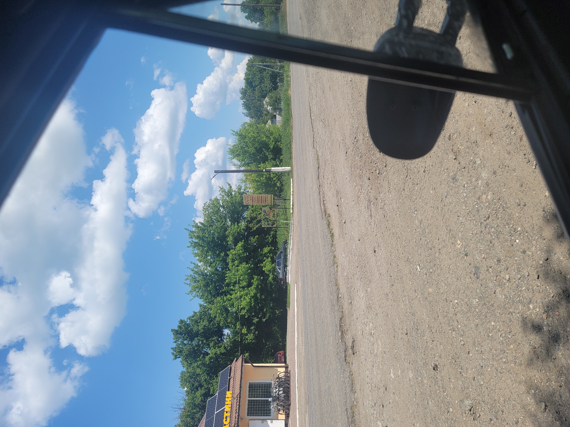 The image depicts a roadside view in Pereshchepyne, Ukraine, on a sunny day. In the foreground, a dirt and gravel road is visible, with shadows suggesting the presence of a vehicle's interior, possibly from the perspective of looking out a window. To the left, a small building with a brown roof and solar panels is situated, with several bicycles parked outside. Behind the building and extending across the midground are lush green trees. Further back, a gray sedan is parked on the side of the road, and other buildings and trees are visible in the distance. The sky is bright blue with scattered white cumulus clouds. A signpost with text is also present to the right of the building.