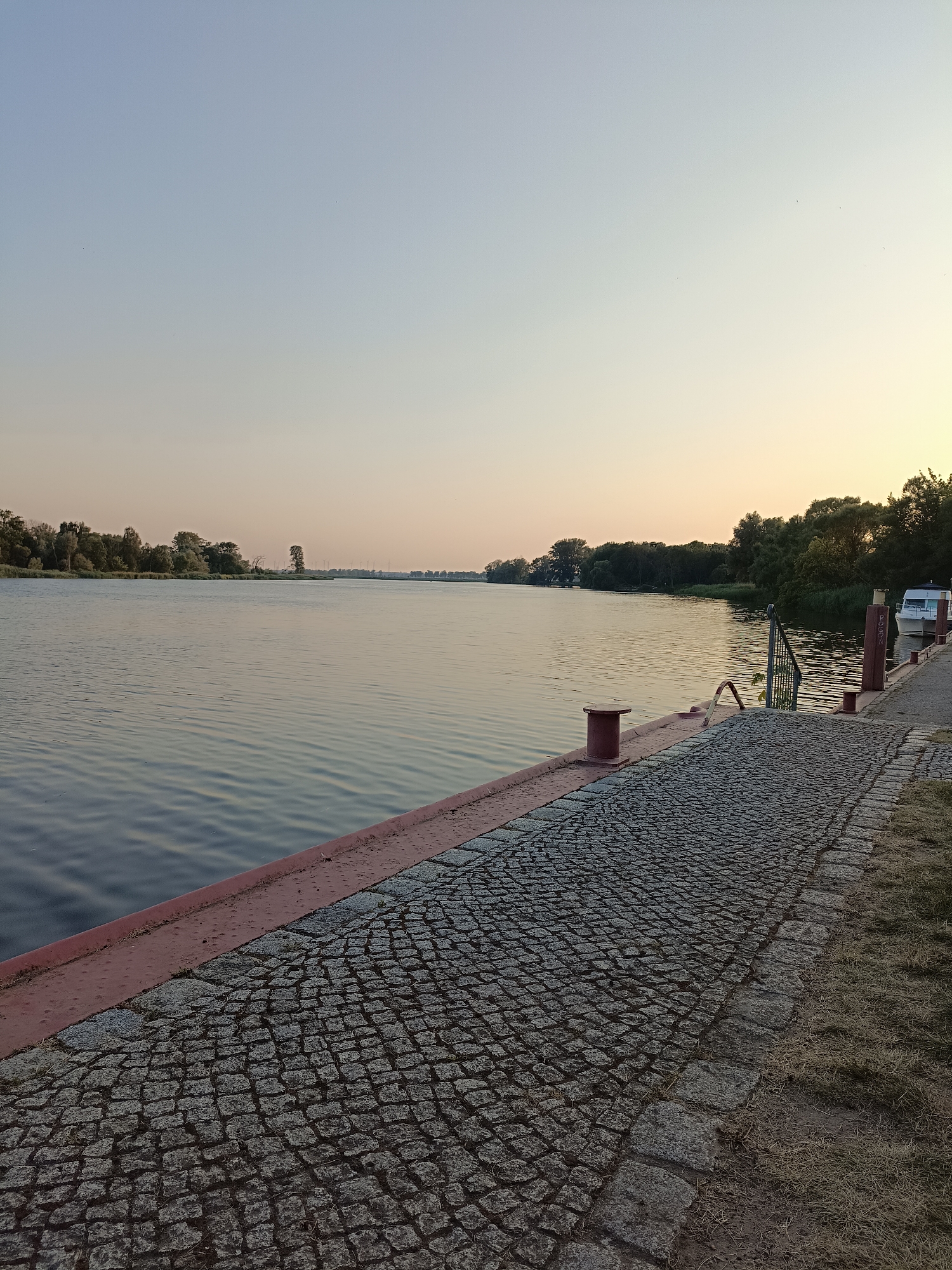 The image depicts a serene scene along the Oder River in Gartz (Oder), Germany, at dusk. The foreground features a cobblestone walkway bordered by a reddish-brown metal railing. The walkway leads to a dock where a small white boat is moored. The river itself is calm, reflecting the soft hues of the twilight sky. Trees and vegetation line the opposite bank, creating a natural backdrop. The overall atmosphere is peaceful and tranquil, suggesting a quiet evening by the water.