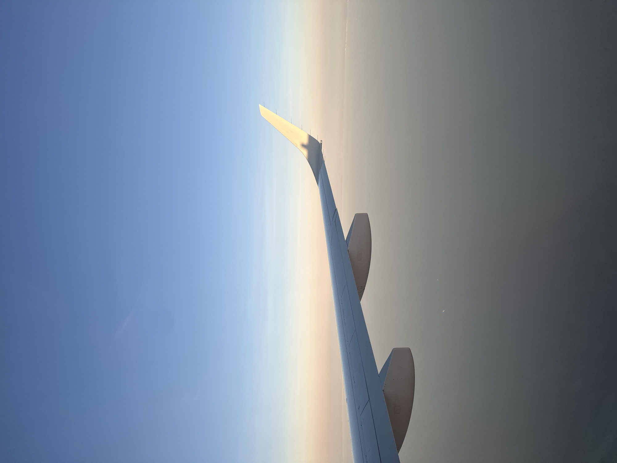 The image depicts an airplane wing viewed from the inside of the aircraft, juxtaposed against a gradient sky. The sky transitions from a light blue at the upper left to a hazy, greyish-brown at the lower right. The wing extends diagonally across the frame, with two flaps visible. The picture was taken outside of Boismont in France.