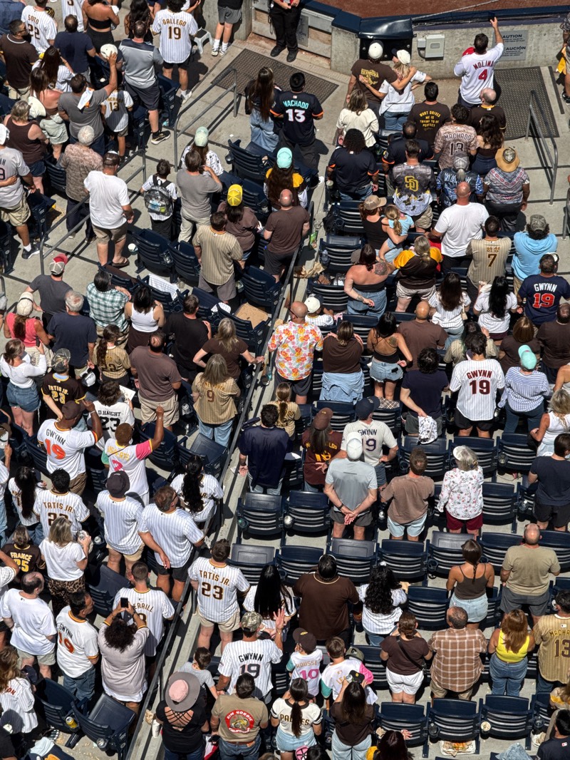 This image shows a large group of people sitting in stadium seats at a baseball game. Most people are wearing baseball jerseys, with many of the jerseys having "GWYNN" and the number 19 on the back. The name "TATIS JR" is also visible on a number of jerseys. Other names include "GALLEGO", "FRANCE", "DHAIGHT", "MOLINI", "MACHADO", "HA-SEONG", and "HOUSSE". The baseball stadium appears to be in San Diego, United States, based on the Padres jerseys.