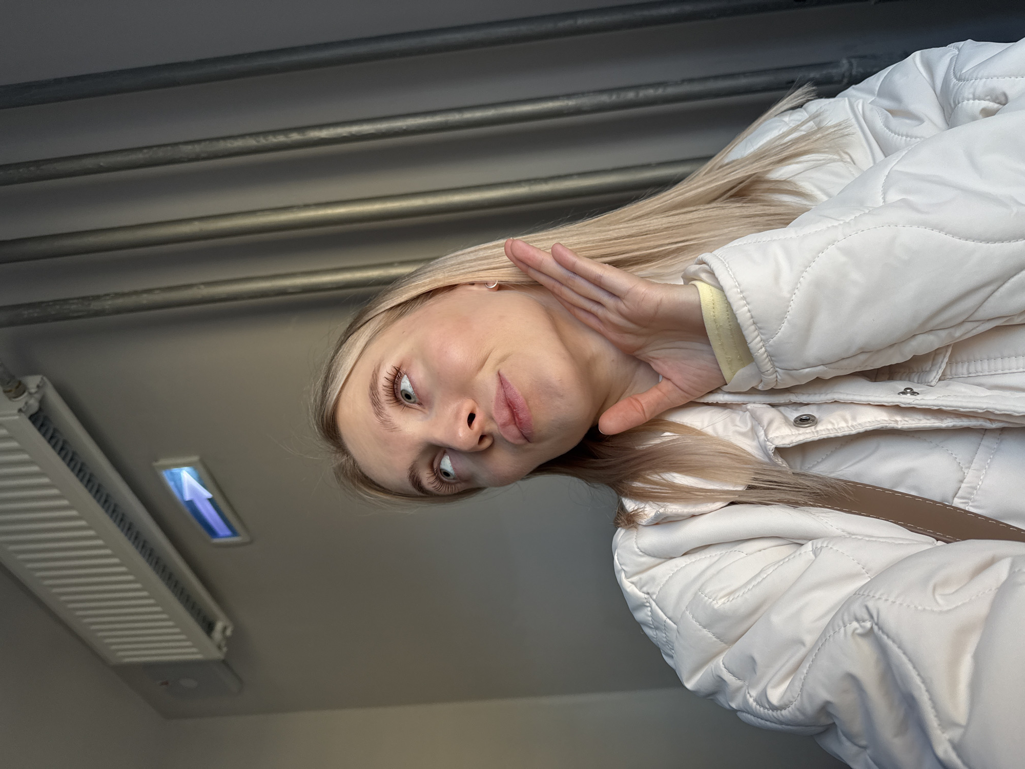 A low-angle shot shows a young, fair-skinned woman with blonde hair wearing a white quilted jacket. She is looking down at the camera with a slightly puckered expression and her hand raised near her face. In the background, there is a grey wall with metal pipes running horizontally. A white radiator with a blue arrow is visible in the upper left corner. This scene is located in Izhevsk, Russia.