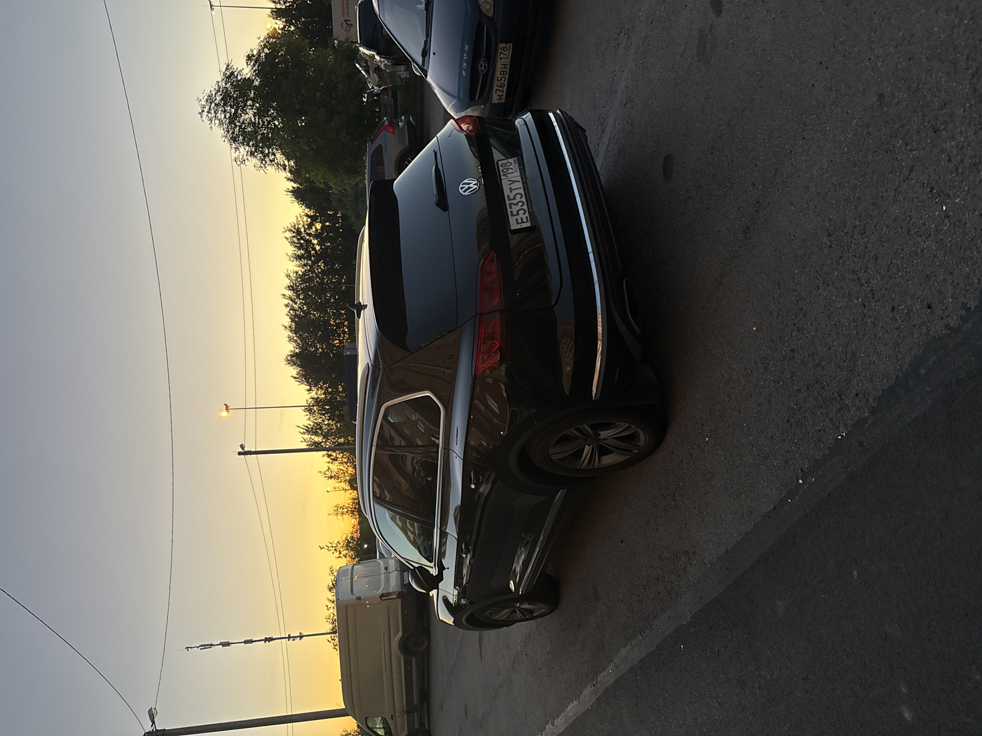A black Volkswagen Touareg is parked on an asphalt road during sunset. The sky is orange and yellow, with a few power lines visible. To the left, a white van is parked, and further in the background, trees and buildings can be seen. A dark-colored hatchback car is parked to the right of the Touareg. The license plate of the Touareg reads "E535TY 198 RUS". The license plate of the hatchback reads "H765BH 178". The scene appears to be a street parking area in Saint Petersburg, Russia.
