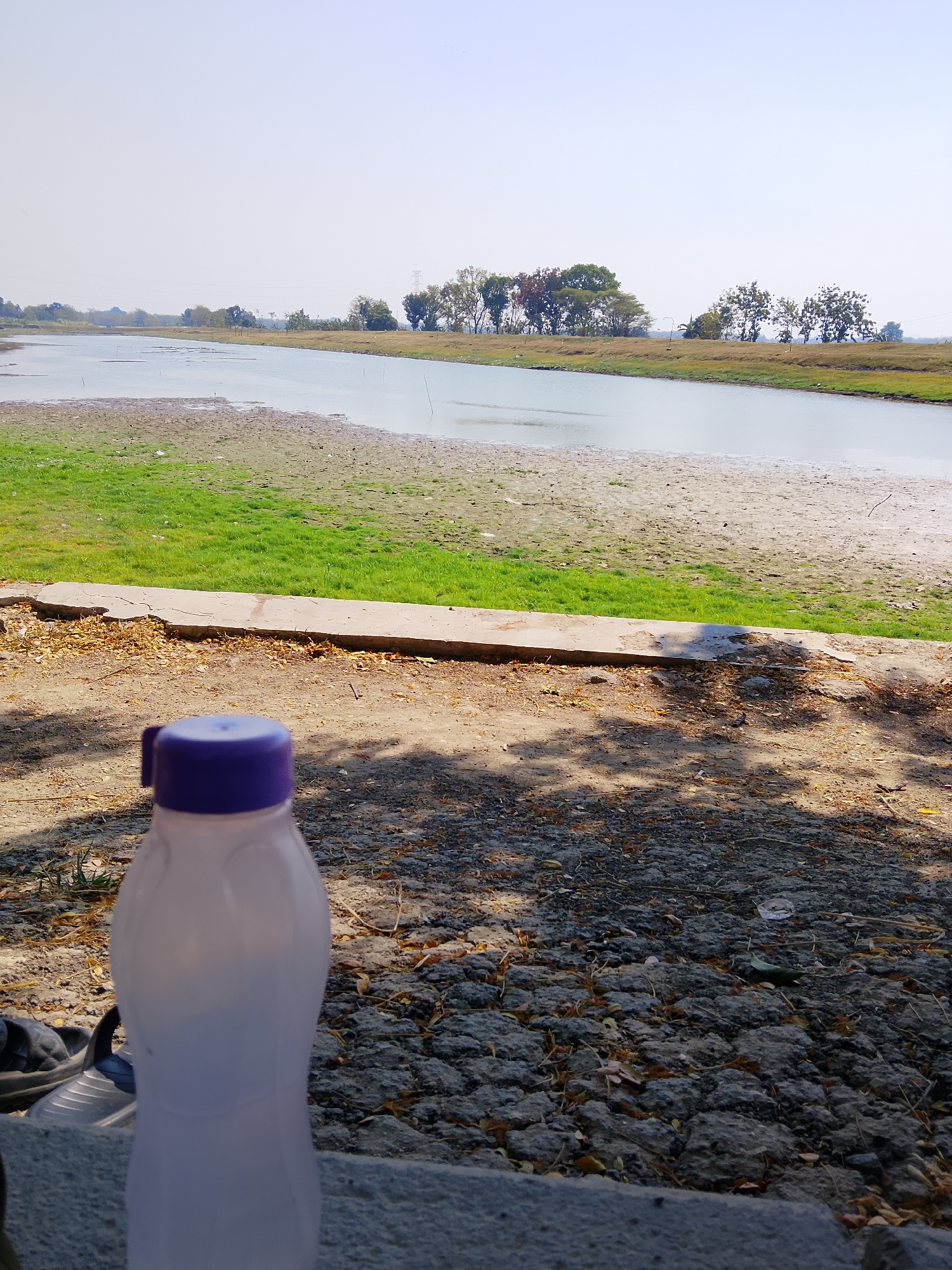 A translucent white plastic water bottle with a purple cap is positioned in the foreground, slightly off-center to the left. It rests on a grey, textured surface scattered with dry leaves and small stones. Behind the bottle, a concrete pathway or ledge is visible, cracked and weathered. Beyond this, a grassy area transitions into a sandy bank that meets a body of water. The water extends into the middle ground, with a distant tree line forming the horizon under a pale, clear sky. The overall scene depicts an outdoor environment, likely near a river or lake, with evidence of natural debris and some man-made elements. The location is identified as Ngadrin, Indonesia.