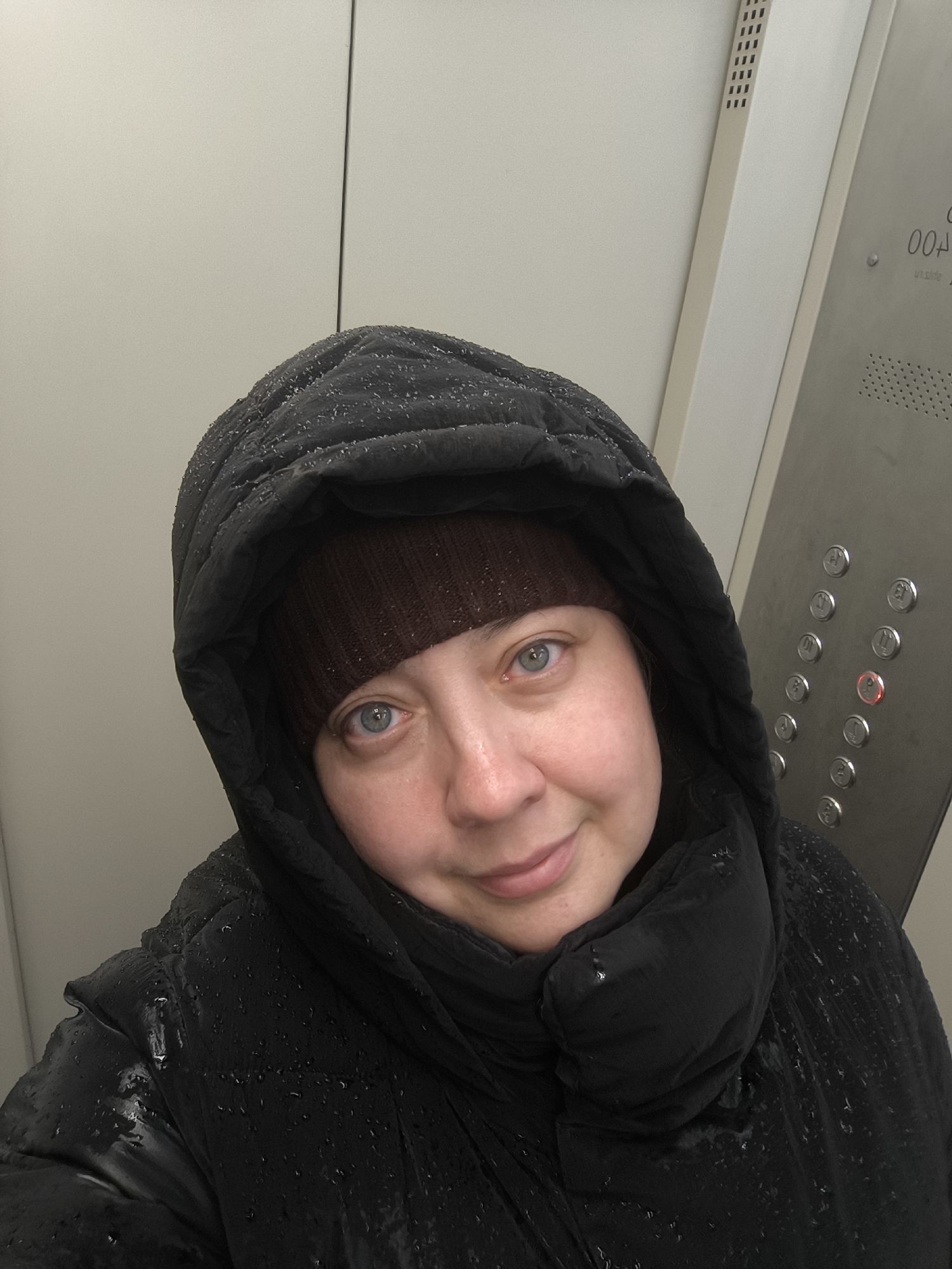 A woman is taking a selfie in what appears to be an elevator. She is wearing a black, possibly waterproof, hooded jacket with what looks like light snow or rain droplets on it. Underneath the hood, she is wearing a dark brown knit hat. Her face is visible; she has fair skin, light blue eyes, and a slight smile. The elevator’s control panel is visible to the right, with round buttons and a red light illuminating what seems to be the ‘P’ button. The elevator's interior walls are a pale gray or off-white color. I can not determine the location as being in the city of Khimki, Russia based on the available image.