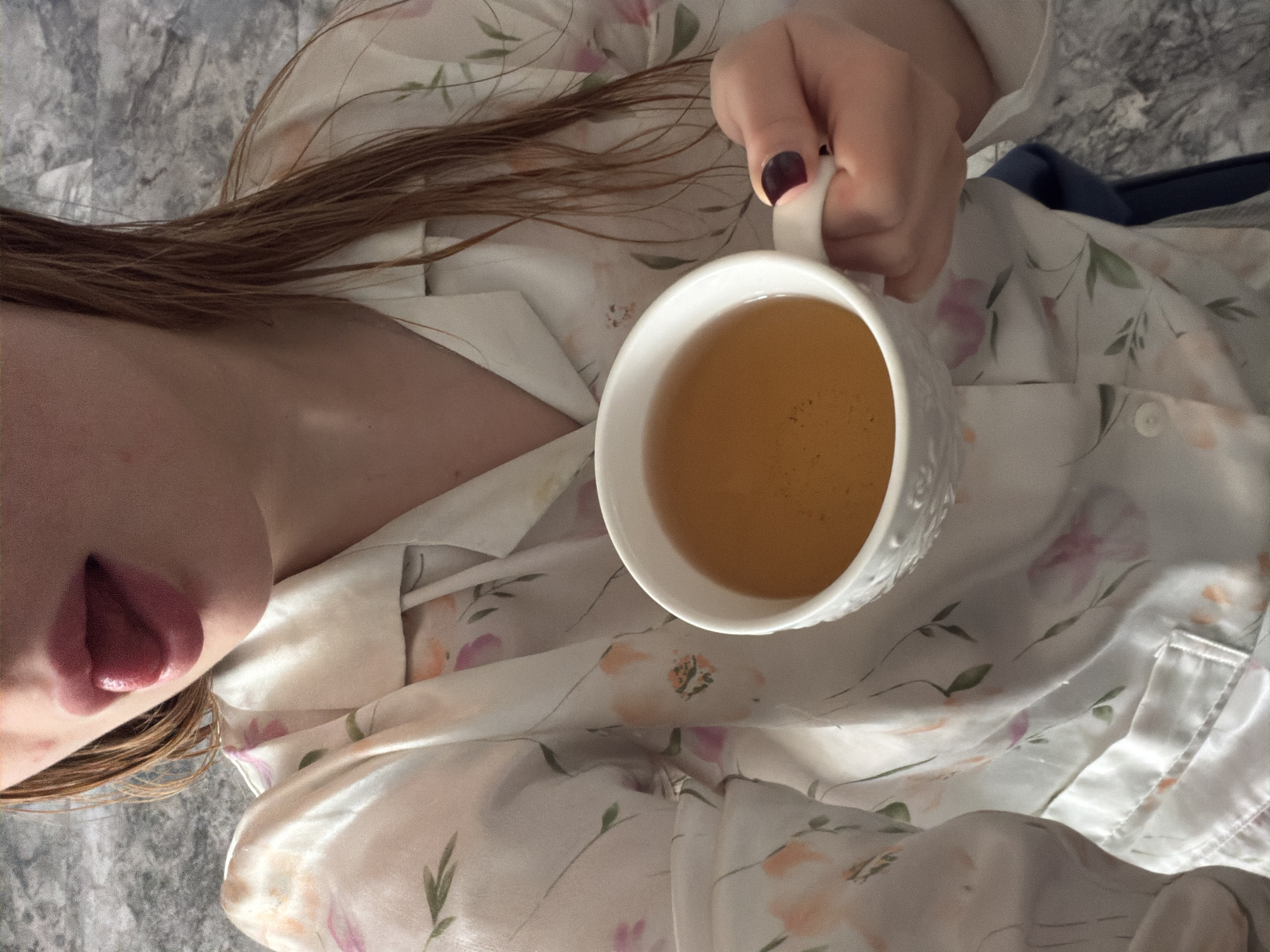 The image shows a person lying down, wearing a floral print shirt, holding a white mug filled with a yellow-brown liquid. The person's face, partially visible, shows lips and hair. A manicured hand with dark nail polish holds the mug. The background is a mottled gray surface, likely a bed or blanket. The exact location in Rossiyskiy, Russia is indeterminable from the image.