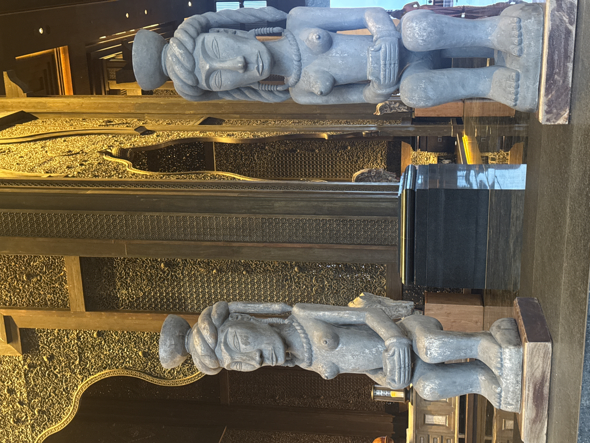 The image shows two identical statues of a seated woman, possibly made of stone or concrete, in an elaborate interior setting. The statues are placed one above the other, and the reflection of the top statue can be seen below the bottom statue. The woman has prominent breasts, braided hair with a headpiece, and holds her hands near her chest. The background features intricate wood carvings and metalwork, suggesting a location of cultural significance, which is Nusa Dua, Indonesia.