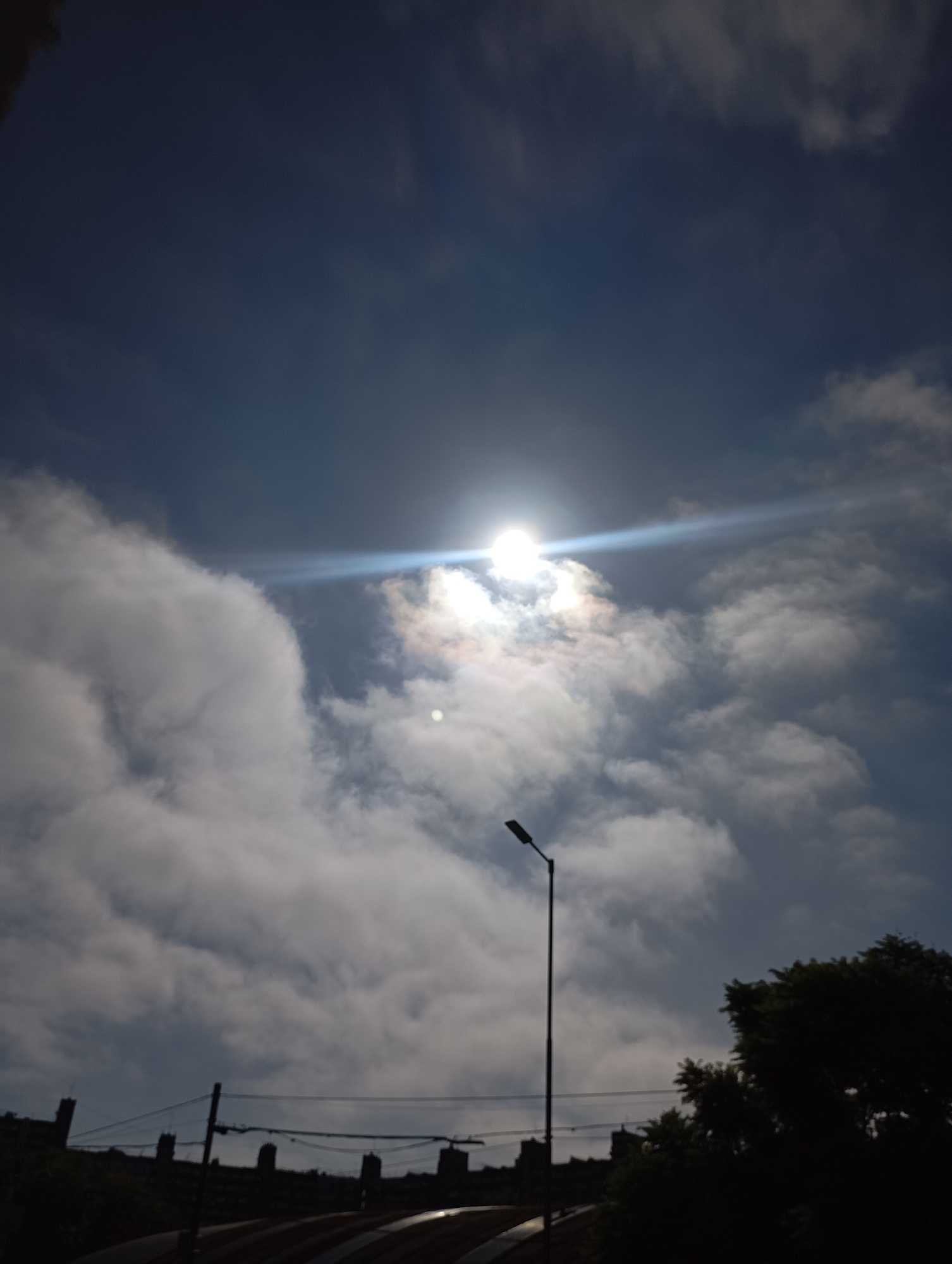 The image captures a daytime scene in Buenos Aires, Argentina. The sky is predominantly blue with scattered, billowy white clouds, partially obscuring the sun. A bright solar flare extends from the sun. A solitary street light stands prominently against the cloudscape. In the lower portion of the frame, silhouettes of buildings and trees are visible, indicative of an urban landscape.