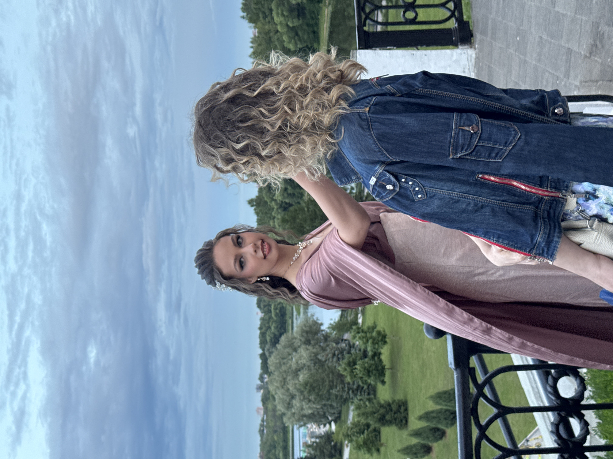 In this vertical, eye-level, medium shot, two young women stand on a balcony with a railing, overlooking a park with green trees and a body of water. The woman in the foreground is wearing a shimmering pink dress with a flowing cape and is adorned with jewelry, including a tiara and earrings. She has long, wavy brown hair and is looking at the camera with a smile. The woman in the background is wearing a denim jacket and has blonde, curly hair. She is turned away from the camera, with her arm around the woman in the pink dress. The sky is cloudy and overcast, suggesting it is daytime. The setting appears to be a park or garden area in Заволжье, Russia, possibly a special event such as a graduation or wedding. The overall mood is celebratory and joyful. There is no visible text in the image.