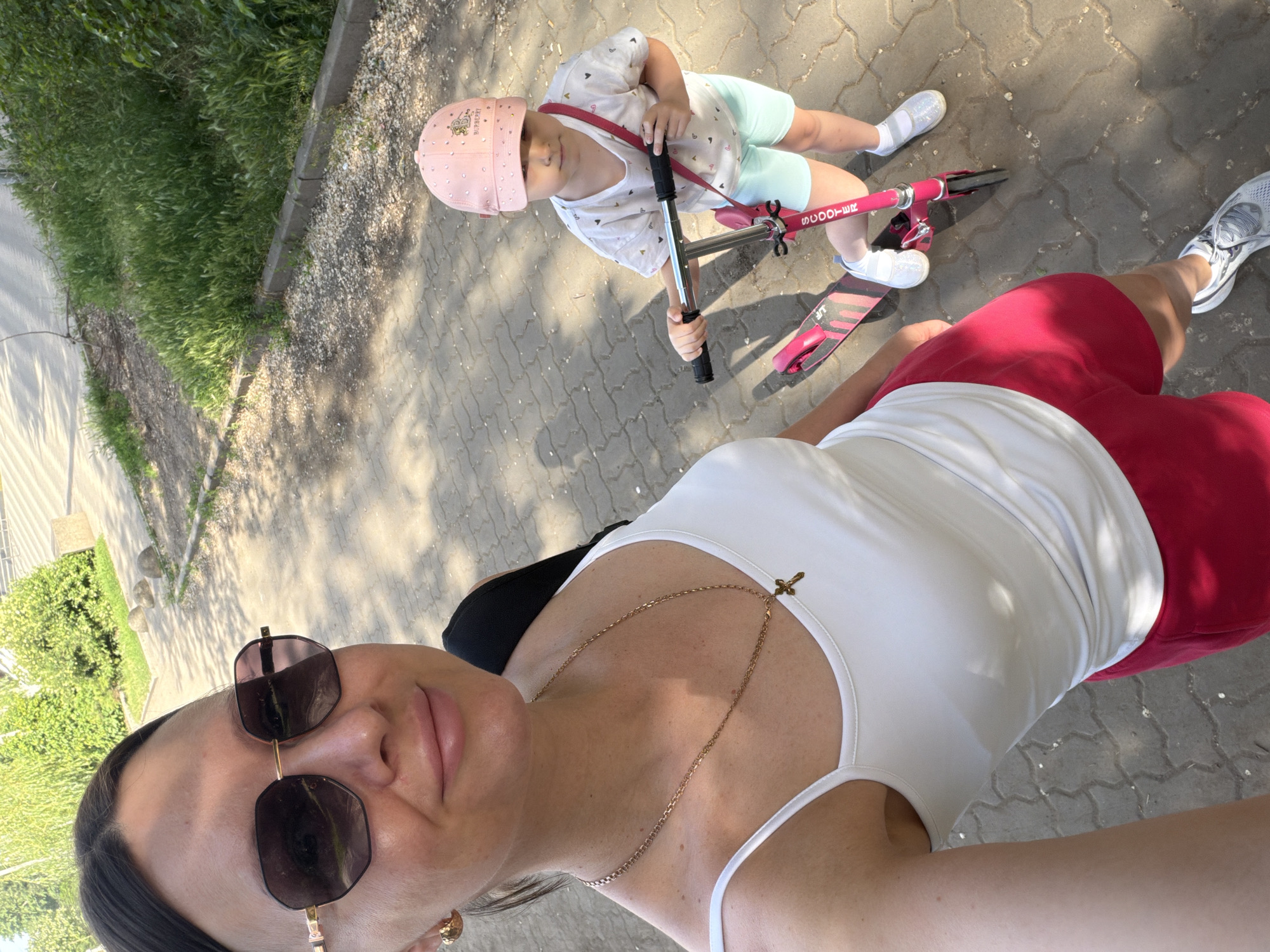 A selfie-style, high-angle outdoor shot shows a woman wearing sunglasses, a white tank top, and red shorts standing on a grey paved surface. She is wearing a gold necklace with a cross pendant. Next to her is a child on a pink scooter, wearing a pink helmet, a white t-shirt with patterns, and light green shorts. Both appear to be on a paved walkway alongside greenery, likely in a city. The environment looks like a park or public space within the city of Rostov-on-Don, Russia.