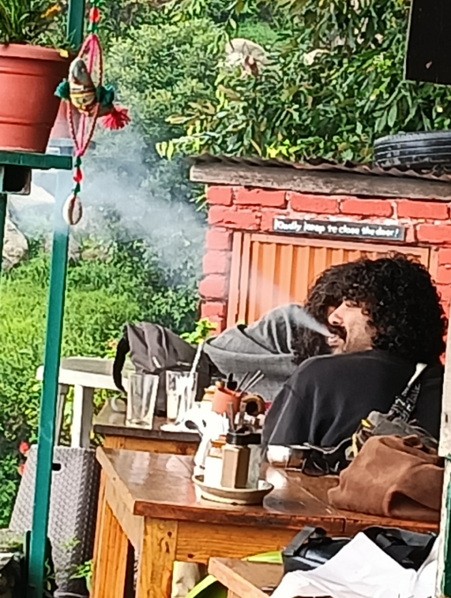 In Haini, India, two individuals are seated at an outdoor wooden table. Smoke emanates from the person on the right, who has dark, curly hair and a mustache, and appears to be smoking or vaping. The person on the left, also with dark curly hair, is wearing a grey sweatshirt. The table is laden with various items, including glasses, a coffee maker, and bags. In the background, a brick wall with a sign that reads "loudly jump to close the door?" is visible, along with lush green foliage and a terracotta pot.
