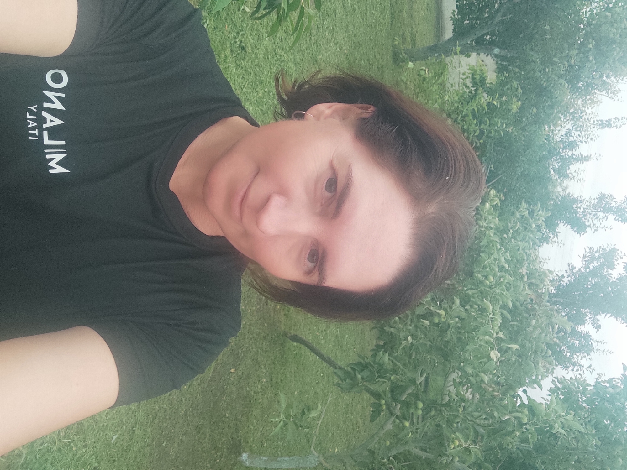 A mid-shot, selfie-style photograph captures a woman with shoulder-length brown hair and light skin. She is wearing a black t-shirt with white lettering that reads "MILANO YJATI". A small silver hoop earring is visible in her left ear. She is smiling slightly and looking directly at the camera. The background consists of a grassy area with several trees and green foliage, suggesting an outdoor setting, likely in Kobeliachok, Ukraine. The lighting is overcast.