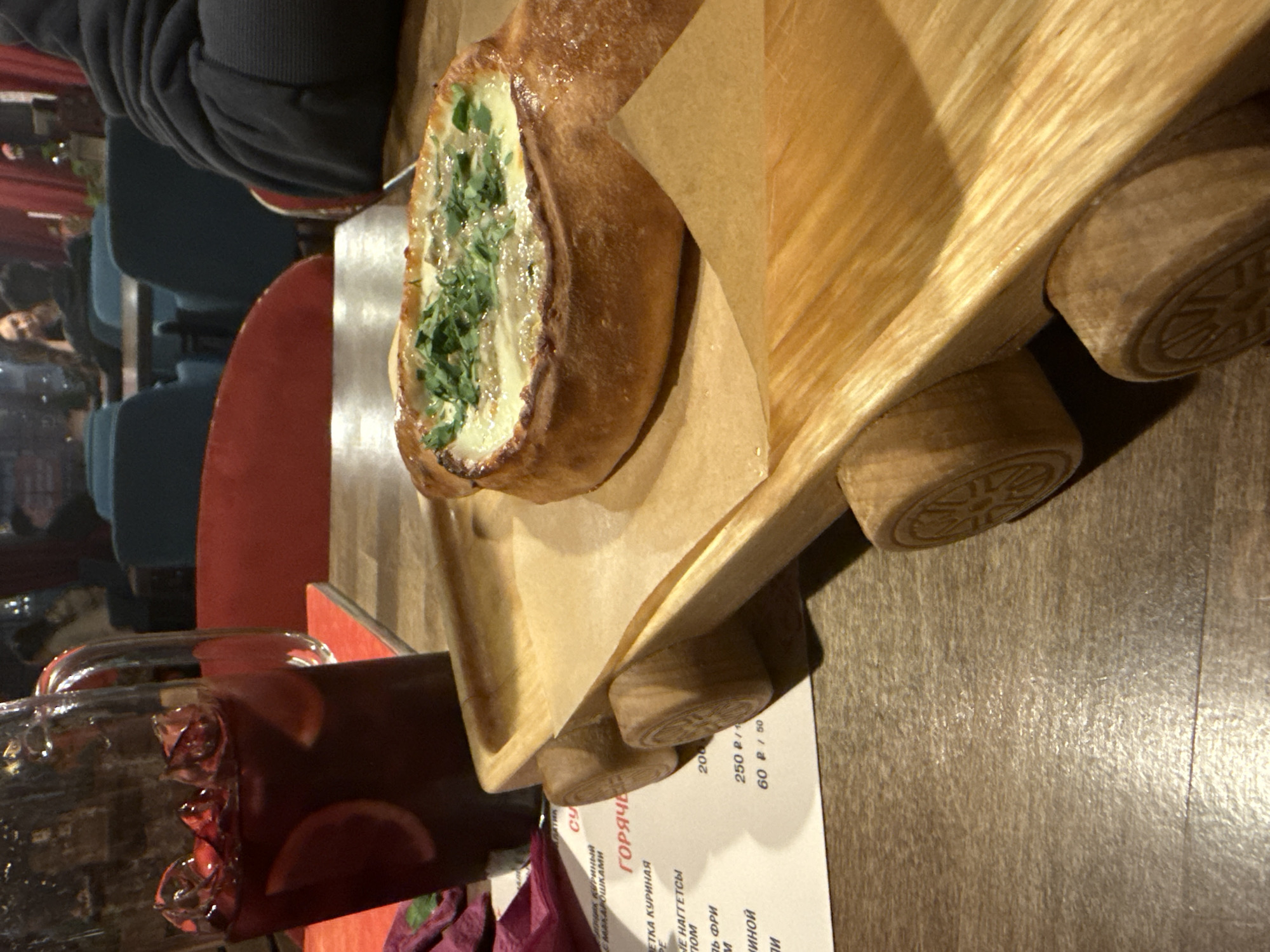 Here is a description of the image:
A close-up shot shows a Khachapuri (Georgian cheese bread) filled with greens, resting on a wooden serving board. The board has a distinctive design with wheels, making it appear like a small cart or train. Next to the board, there is a glass pitcher of a dark-colored beverage containing sliced fruit, likely sangria. Part of a menu with Russian text is visible beneath the board, with items and prices listed. The background shows a blurred indoor scene with people and chairs, suggesting a restaurant or cafe setting.
Based on the visible text in the menu (translated), "ГОРЯЧЕ (Goryache)" means "HOT," indicating this photo was taken in a Russian-speaking environment. The currency listed on the menu is Rubles, and judging by the image content and location, it is highly possible the restaurant is located in Perm, Russia.