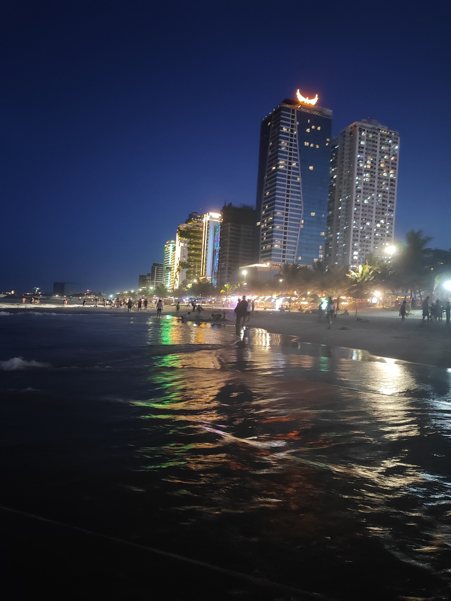 This nighttime scene depicts a beach in Đà Nẵng, Vietnam. A row of brightly lit high-rise buildings lines the coast. The foreground shows a dark, reflective body of water, presumably the sea, with colored light reflections. On the sandy beach, people are walking, standing, and interacting, mostly in silhouettes. A few palm trees are visible along the beach. The sky is a deep blue, darker than the illuminated coastline. The buildings have a variety of lights, including a distinct crescent-shaped light atop one of the tallest structures.