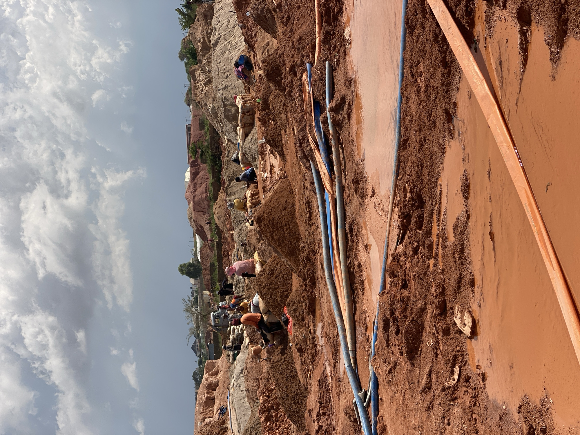 The image depicts a group of people working in a muddy, reddish-brown terrain, likely a construction or excavation site. The scene is oriented vertically, suggesting the original photo was taken sideways. The subjects are individuals engaged in manual labor, some appearing to be digging or moving earth. Several are dressed in casual clothing, with one person wearing a pink garment.
Objects in the scene include numerous pipes of varying colors, predominantly blue and orange, lying on the ground. Some pipes are submerged in the muddy water. The ground itself is a mixture of soil, rocks, and water, indicating a wet and possibly recently rained-upon environment. In the background, a rocky hillside rises, with some vegetation and buildings visible at the top, suggesting an inhabited area or a nearby town. The sky is partly cloudy, with patches of blue visible.
The activity appears to be related to earthmoving or construction work, possibly involving water management due to the presence of standing water and pipes. The time of day seems to be daytime, given the illumination and the sky conditions. The location cues, such as the reddish soil and the style of buildings in the background, align with the provided context of Reifild-Ist, Nigeria. The overall impression is one of arduous manual labor in a challenging environment. No visible text is present in the image.