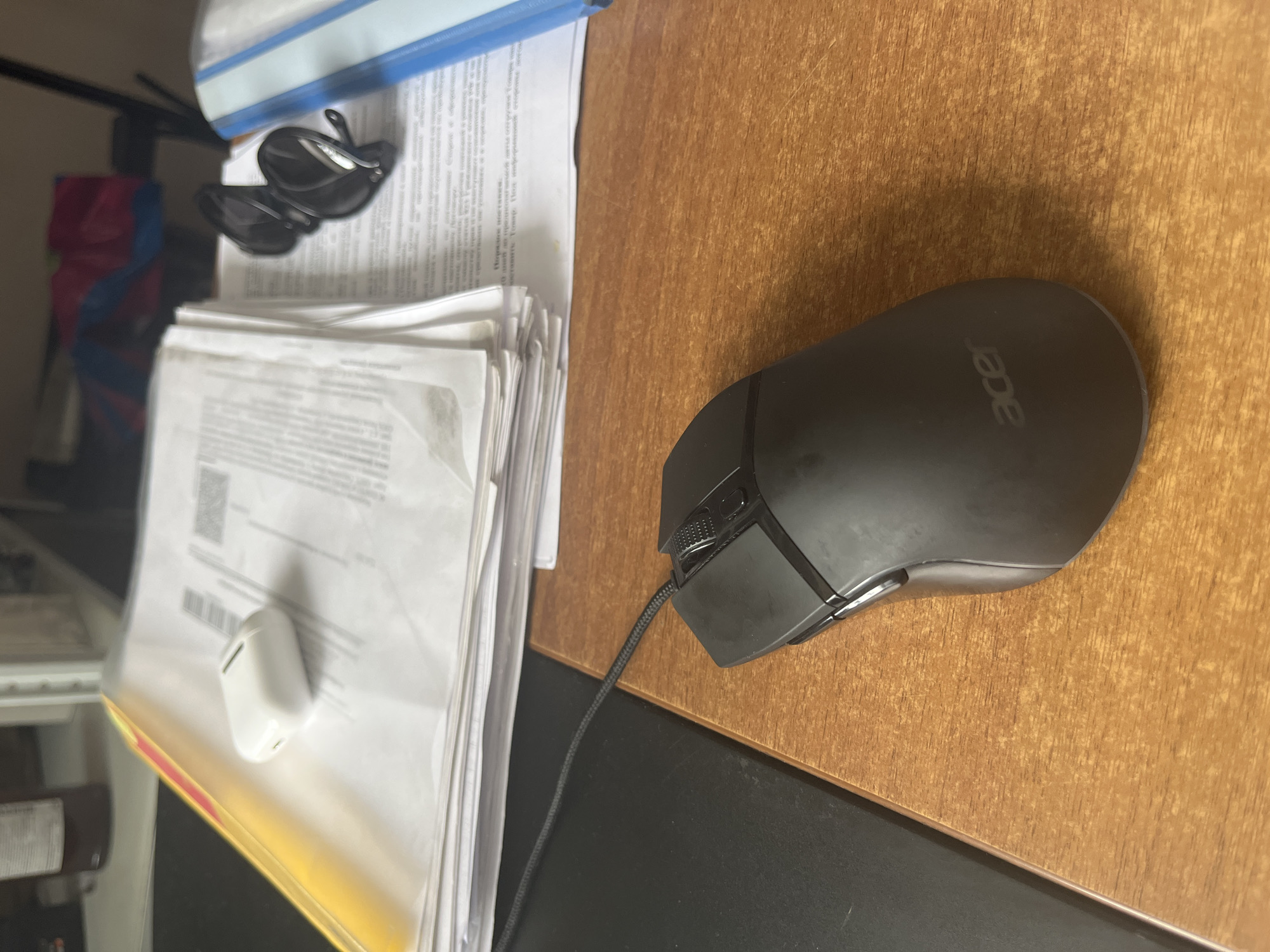 The image depicts a close-up, high-angle view of a wooden desk surface with various office-related items. In the foreground, a black wired computer mouse with an "acer" logo is positioned to the right. Behind the mouse, a stack of white papers is visible, partially obscured by a yellow folder on the left. A white charging case for wireless earbuds rests on top of the papers. To the right of the papers, a pair of black sunglasses and a blue and white binder are also present. The background is blurred, showing what appears to be shelving and a dark object, possibly a bottle. The overall setting suggests a workspace. The image does not contain any information that would allow for the determination of its location in Yekaterinburg, Russia, or any other specific geographic area.