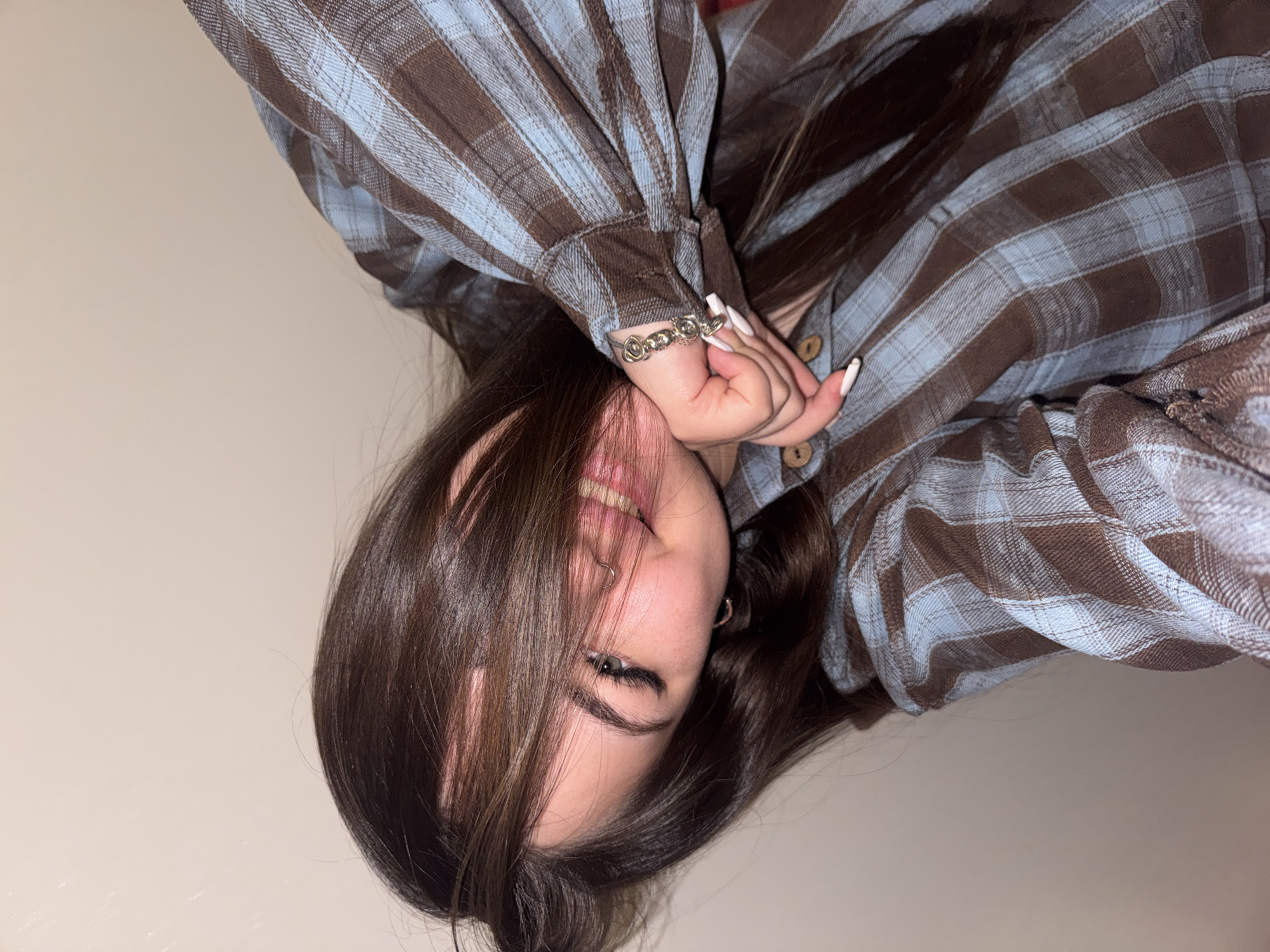A young female with long, dark brown hair is captured in a close-up, sideways-oriented portrait, likely taken indoors in Murino, Russia. She is smiling broadly, showing her teeth, with some of her hair falling playfully over her face. Her left hand is raised towards her face, revealing long, white, pointed fingernails and a silver-toned bracelet featuring an intricate, intertwined design, possibly roses or hearts.
She is wearing a long-sleeved plaid shirt with a pattern of brown and light blue checks. The shirt features wooden buttons on the placket and sleeve cuffs. A silver-toned hoop piercing is visible in her left nostril, and a small silver-toned hoop earring can be seen in her right ear. Her visible eye shows a neatly shaped eyebrow and dark eyeliner. The background is a plain, light-colored wall, indicating a simple interior setting. No text is visible in the image. The overall impression is one of a cheerful and casual moment.