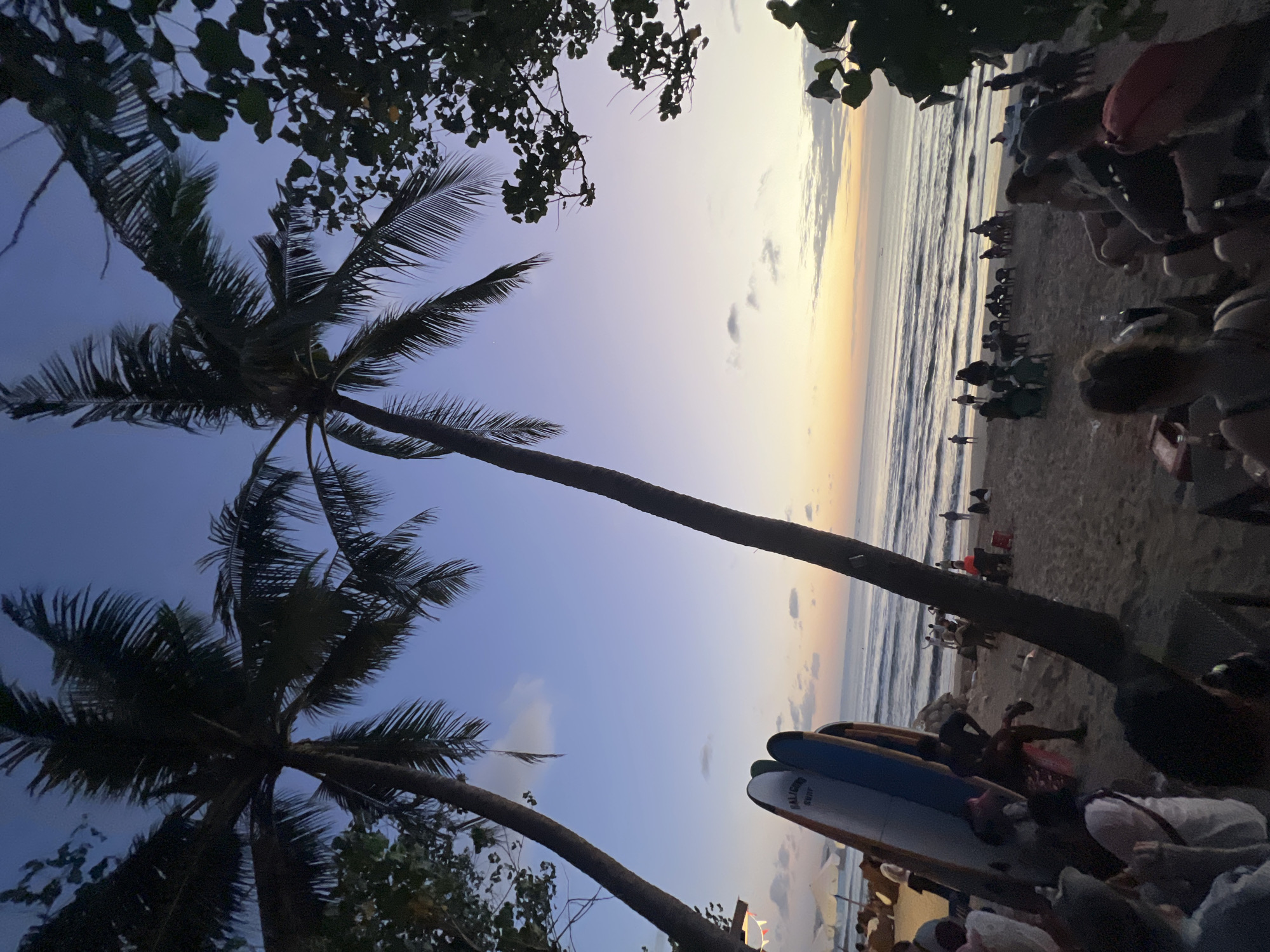 In Kuta, Indonesia, at dusk, two silhouetted palm trees dominate the foreground, their fronds creating a textured canopy against a twilight sky that transitions from deep blue overhead to pastel shades of purple, pink, and orange near the horizon. The scene unfolds on a sandy beach with gentle waves lapping at the shore. Several surfboards, one with "BALICULT SURF" visible, stand upright on the sand. A group of people are scattered across the beach, some seated in chairs, others standing and walking. In the mid-ground, figures are gathered in small groups, some appearing to be dining or socializing at tables. In the background, the ocean stretches to the horizon, with a faint boat visible. The overall atmosphere is relaxed and serene, capturing the end of a day at a popular Indonesian beach destination.