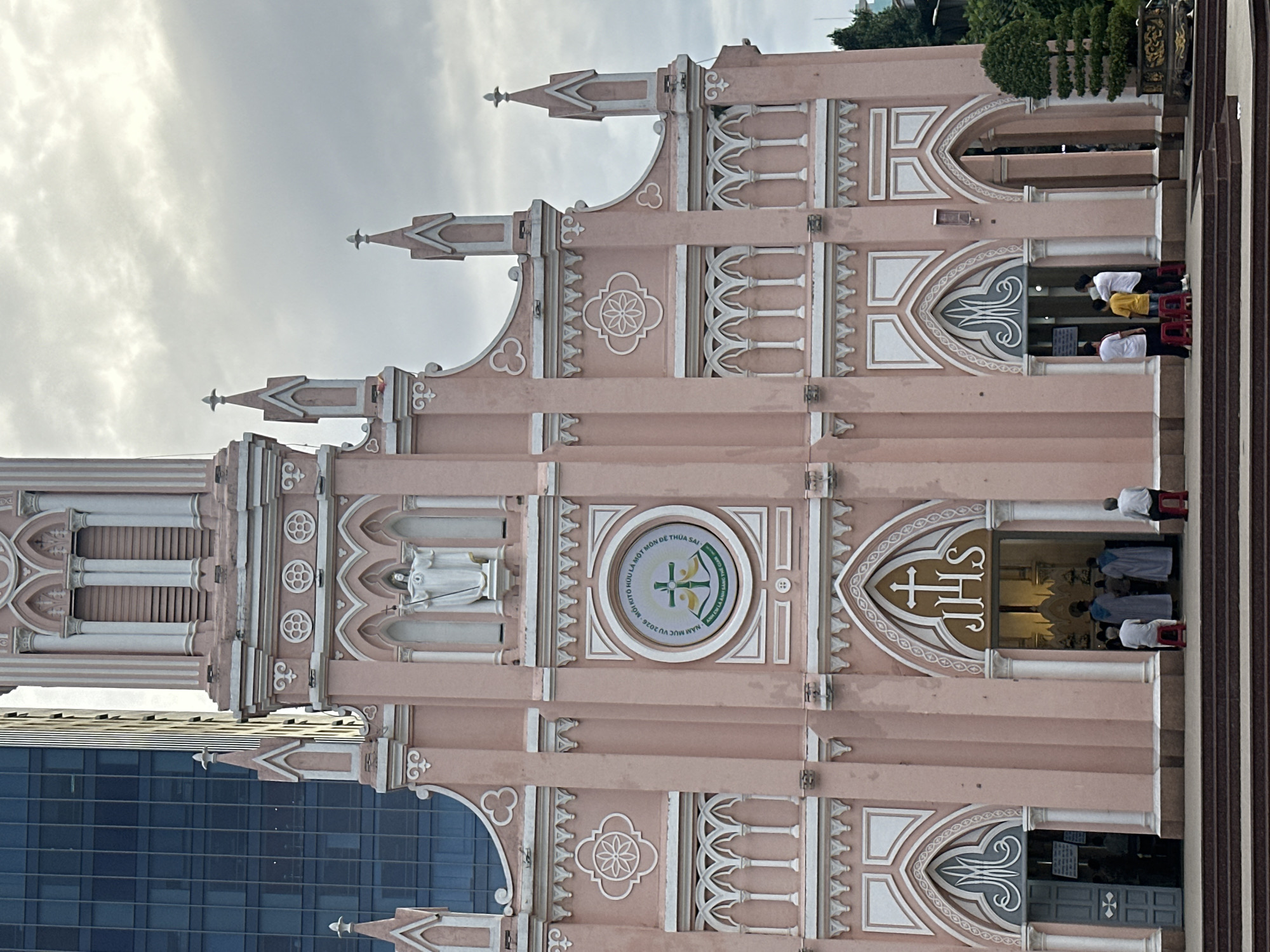 The image shows the pink facade of the Da Nang Cathedral, also known as the Church of the Sacred Heart of Jesus, located in Da Nang, Vietnam. The Gothic Revival-style church is painted a vibrant pink with white trim and features a tall, slender tower. In front of the entrance, there are several people, some standing and some sitting on red stools. One entrance has a gray door with a cross symbol. Above the main entrance, a circular emblem with a cross and text is visible. A modern glass building looms behind the church on the left. The sky is overcast with clouds.