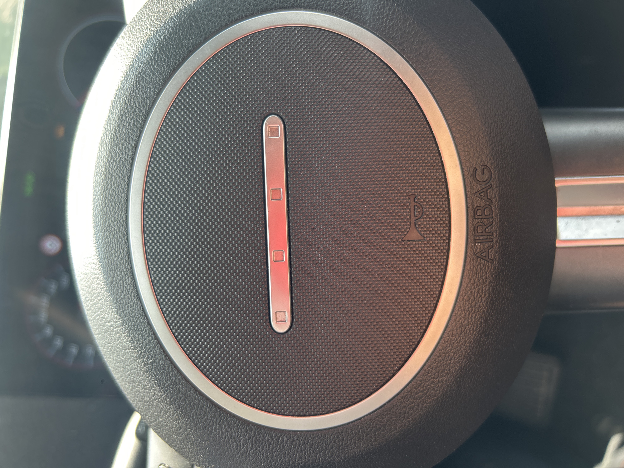 The image provides a close-up, eye-level view of the center of a car's steering wheel. The primary subjects are the steering wheel itself and its central airbag cover. The steering wheel is dark gray or black with a textured surface, and the central part is a circular, finely dimpled dark gray panel. This panel is framed by a sleek silver-colored trim ring.
A prominent feature is a vertical, elongated silver strip located in the center of the circular panel. This strip contains three small, square-shaped segments, all of which are brightly illuminated with a red light. To the right of this strip, on the dimpled surface, there's a small, embossed symbol resembling a horn or sound waves. Below this, on the outer rim of the steering wheel, the word "AIRBAG" is embossed vertically in uppercase letters.
The setting is inside a car, likely during daytime, as indicated by bright reflections on the silver trim and the top-left portion of the steering wheel, possibly from sunlight. The background, to the left, shows blurred parts of the car's dashboard or instrument cluster with some indistinct lights. No activity or event is taking place; the image focuses on the static details of the car's interior. No people are visible.