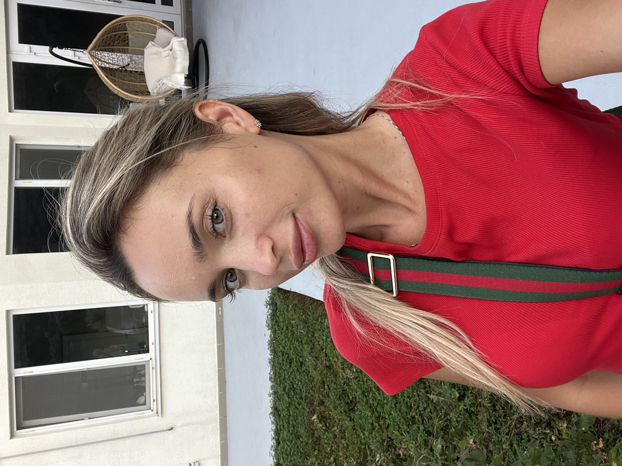 A woman with blonde hair and green eyes poses for a selfie outdoors. She is wearing a red ribbed t-shirt with a green and red striped strap. In the background, there is a white building with large windows, a green grassy area, and a hanging chair. The location is identified as Fontanka, Ukraine.