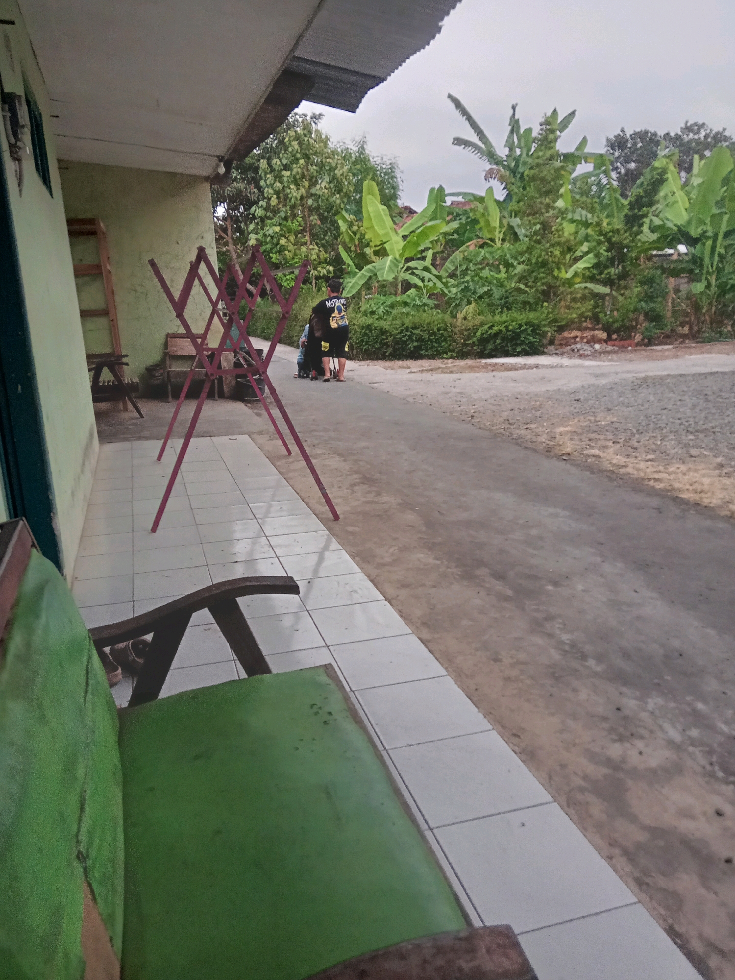 In Desa Klahang, Indonesia, a low-angle shot captures an outdoor scene with a porch and a pathway. On the porch, a wooden bench with worn green upholstery is visible in the foreground. To the left, a light green wall of a building stands with a door and a ladder leaning against it. A pink A-frame clothes drying rack is positioned on the tiled porch.
Beyond the porch, a paved pathway or road runs horizontally across the frame, bordered by lush green vegetation. In the background, several people are gathered near a motorcycle. One individual, wearing a black t-shirt with a graphic and dark shorts, is seated on the motorcycle. Another person is standing beside them. The environment is tropical, with numerous banana trees and other dense foliage evident. The sky appears overcast.