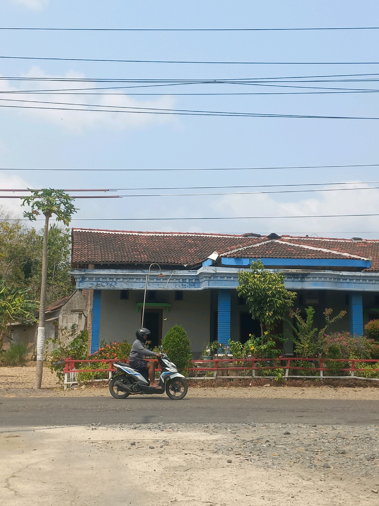 A man wearing a helmet and a grey long-sleeved shirt is riding a white and blue motorcycle on a paved road. He is moving from left to right in the frame. Behind him, a house with a red tiled roof and blue accents on its facade is visible. The house has several windows and a porch supported by blue columns. A red fence runs along the front of the property, in front of various green bushes and flowering plants. To the left of the house, a tall, slender tree with green foliage at its top stands beside the road. The environment appears to be a rural or suburban area, with power lines stretching across the sky, which is partly cloudy. The ground in the foreground is rough and unpaved, with gravel and dirt visible. The scene is captured during daylight.