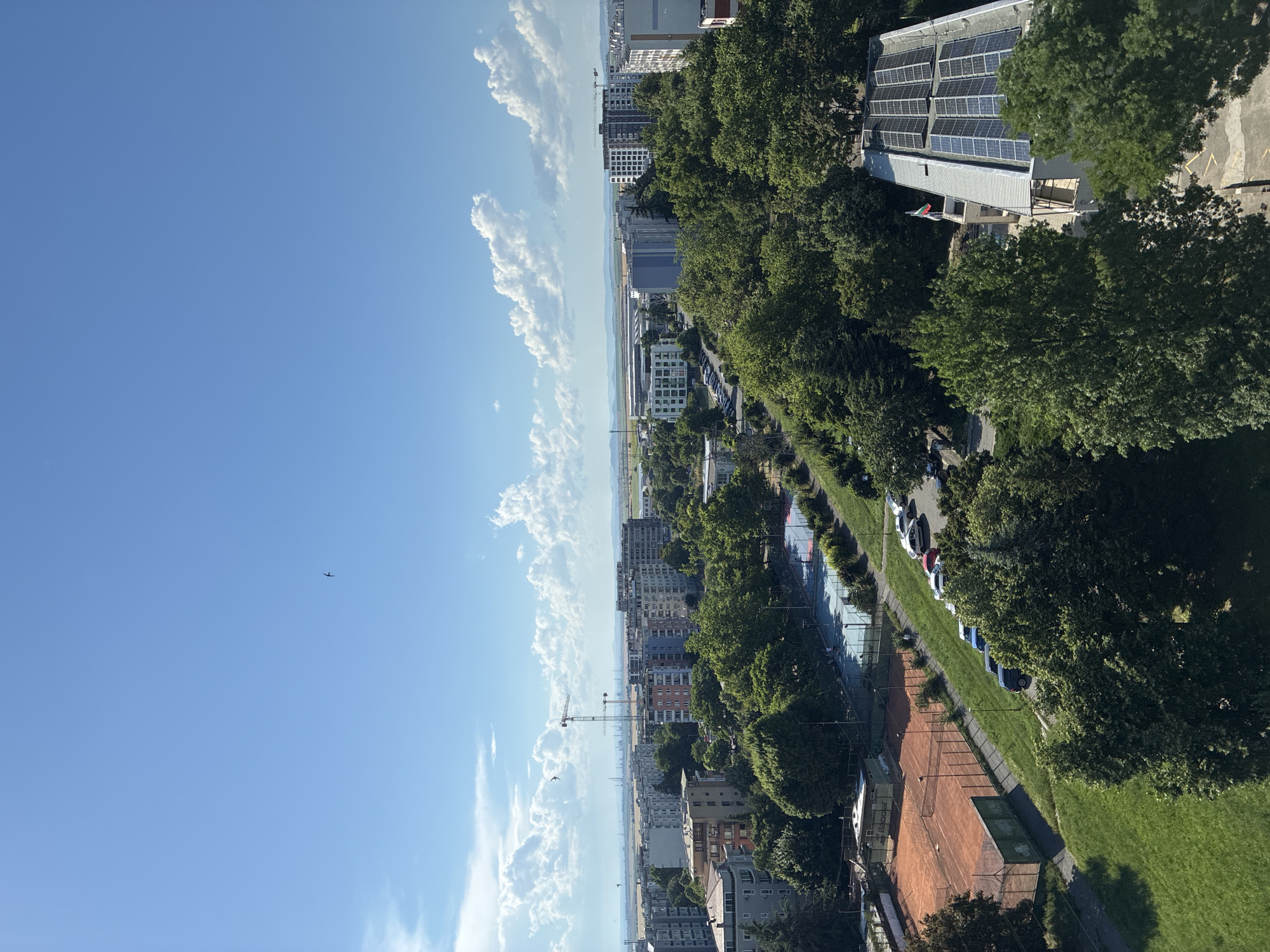 This image depicts an aerial view of the southern part of Burgas, Bulgaria, looking northeast. In the foreground, a green lawn slopes down towards two tennis courts, one clay and one hard surface. Beyond the courts, a street is lined with parked cars and mature trees. Further in the background, a cluster of residential buildings, some with colorful facades, are interspersed with more trees and green spaces. In the distance, industrial facilities with cranes and smokestacks are visible along the coastline, with the sea and a hazy horizon completing the scene under a bright blue sky dotted with fluffy white clouds. A single bird is captured in flight in the upper portion of the sky. A building with a pitched roof covered in solar panels is visible in the lower right quadrant.