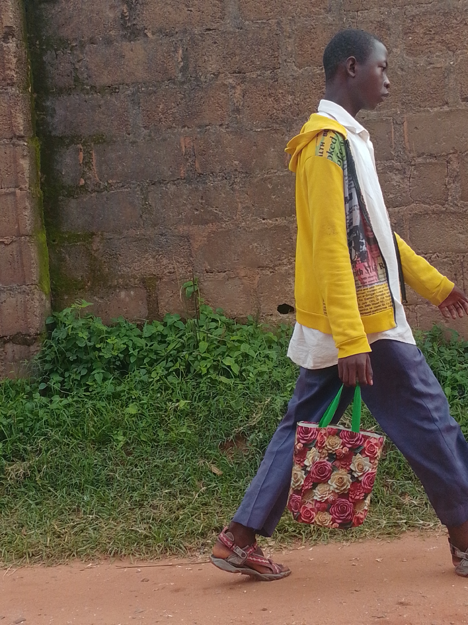 A young Black boy walks from right to left in front of a brick wall, with green grass and foliage in the middle ground and a dirt path in the foreground. He is wearing a yellow hooded jacket over a white shirt with graphics on it, blue pants, and brown sandals. He is carrying a floral-patterned tote bag with green straps in his right hand. The boy is looking to his right. The brick wall is made of reddish-brown bricks, with some green moss growing on it in the upper left corner. The grass is lush and green, with some taller plants interspersed. The dirt path is reddish-brown.
