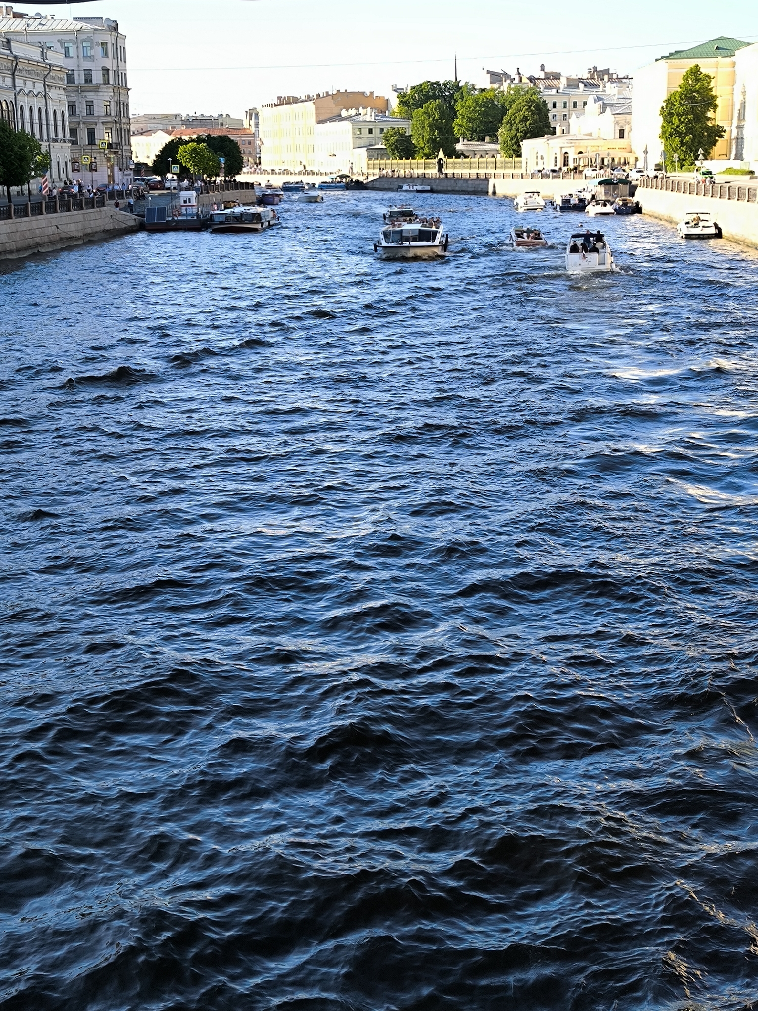 The image captures a detailed view of a bustling urban waterway in Saint Petersburg, Russia, under clear daylight conditions. The lower two-thirds of the frame are dominated by dark blue water, agitated by numerous ripples and small waves, reflecting sunlight in scattered highlights.
The midground reveals two stone embankments lining the waterway. The left embankment features a paved walkway with multiple pedestrians. This walkway is bordered by a stone balustrade and railings. Behind it, a multi-story building of classical European architecture, primarily white or light grey, displays arched windows and ornate detailing. Deciduous trees with green foliage are present along the street, and two yellow diamond-shaped road signs, each bearing a black "A," are affixed to poles. Two rectangular, flat-topped structures, possibly pontoons or small dockside facilities, are moored along this bank.
The right embankment is similarly constructed with stone, a pedestrian walkway, and railings, with multiple people visible. A large, light-colored, multi-story building, potentially yellowish or off-white, with a prominent green roof, stands on this side, also flanked by green trees.
The waterway itself contains a minimum of eight powerboats actively navigating the water. One large, white passenger boat, carrying numerous individuals, is positioned in the left midground and appears to be moving towards the viewer. The remaining boats are smaller, varying from compact motorboats to medium-sized open-top tourist vessels, dispersed across the waterway. Most of these boats are oriented away from or across the camera's perspective. Passengers are discernible within the majority of these moving vessels.
The background presents a dense urban landscape typical of Saint Petersburg, comprising numerous multi-story buildings of varying architectural styles and hues, interspersed with additional green trees. The sky above is clear and bright, indicating sunny weather and good visibility.