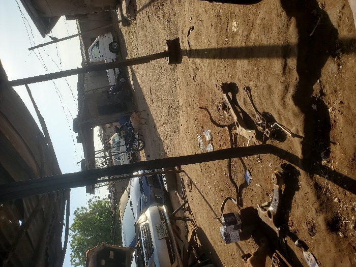 The image displays an outdoor automotive repair area in Kaduna South, Nigeria, under clear, bright daylight. In the left foreground, a silver sedan with an open hood rests on a jack stand, positioned ahead of a dark grey sedan also featuring an open hood. The unpaved dirt ground in the foreground and midground is littered with various discarded automotive components, including suspension control arms and linkages, alongside two white boxed items. Overhead, a rudimentary canopy structure, supported by dark metal poles, provides partial shade. In the midground, a black motorcycle is parked near a white mini-pickup truck, behind which another white vehicle is partially visible. Two individuals are seated or crouching in the midground, near the motorcycle and truck. A light-colored goat walks rightward across the midground. The background consists of several single-story structures appearing to be constructed from concrete or mud bricks.