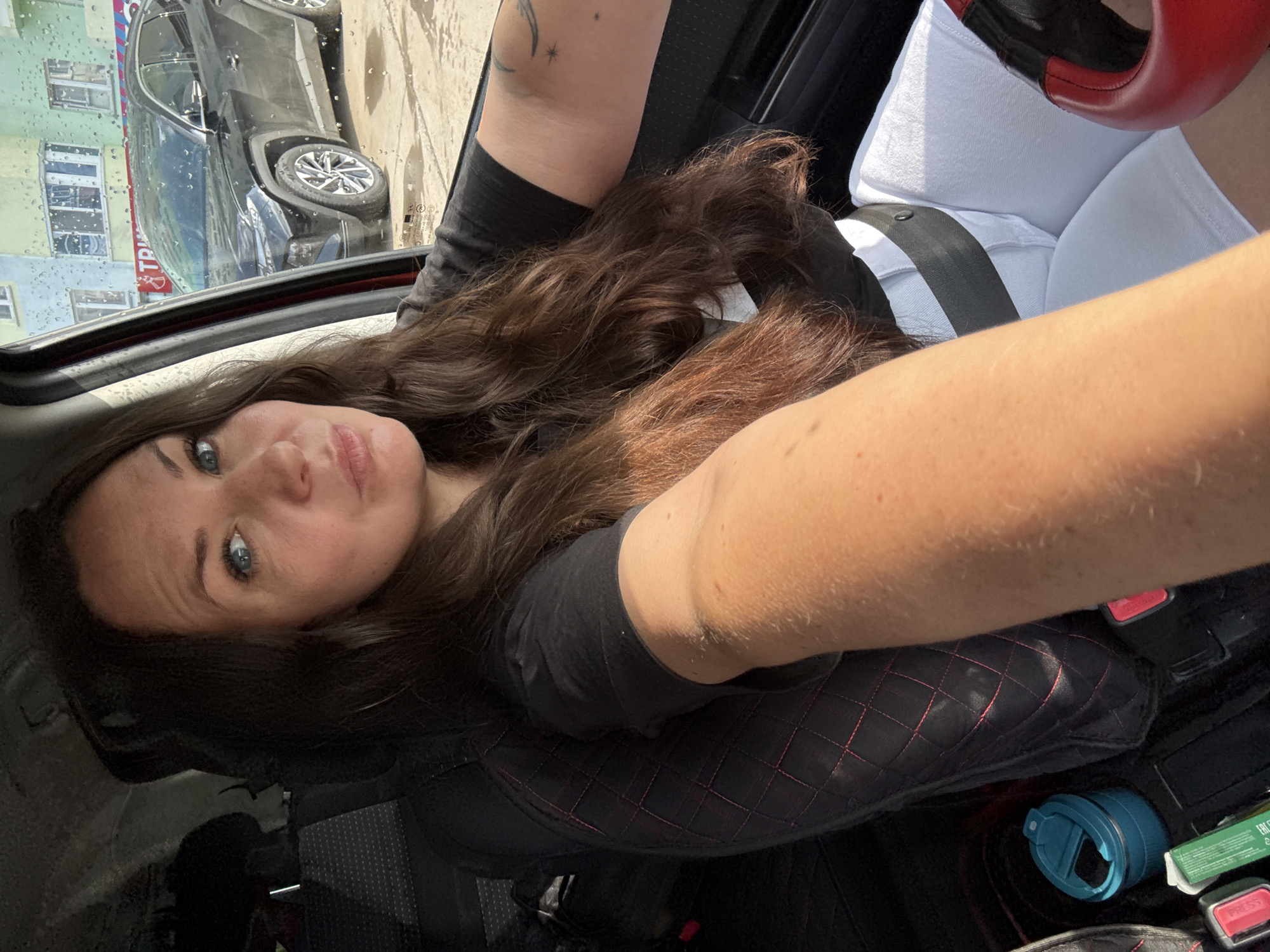 A woman with long brown hair and blue eyes is sitting in the driver's seat of a car. She is wearing a black t-shirt and white shorts, and her seatbelt is fastened. Her left arm is extended forward, and her right arm is resting on the steering wheel. Outside the car, a black SUV is parked on the street in front of a building with colorful walls. Raindrops are visible on the car window. The scene appears to be in the city of Chaikovsky, Russia.