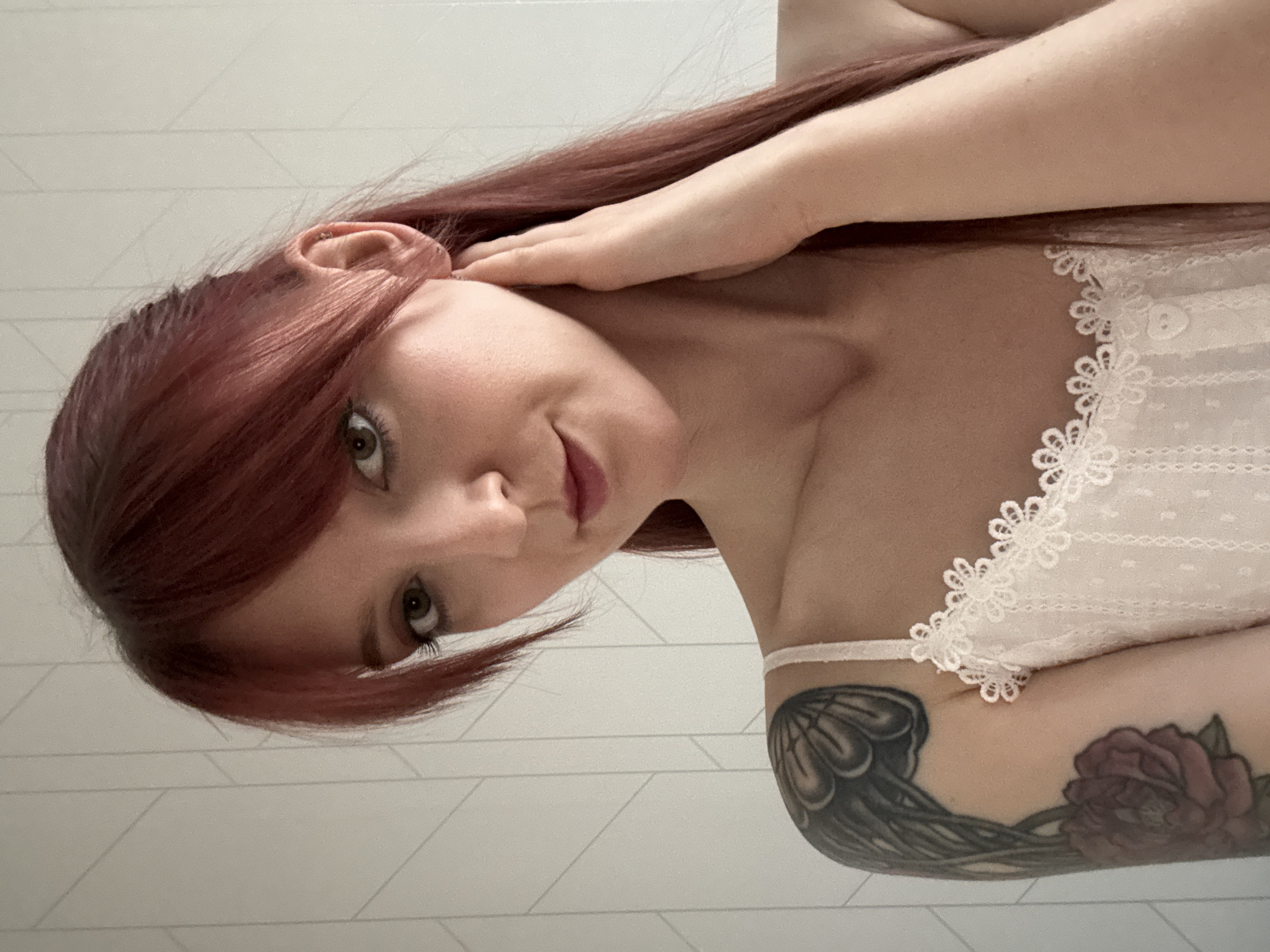 A woman with red hair is shown from a low angle, with her head tilted to the left. She is wearing a white top with a lace trim. Her left arm is raised, with her hand near her ear. A tattoo is visible on her left arm. The background is a white wall. The location is not discernable in the image, and therefore cannot be located in Izhevsk, Russia.