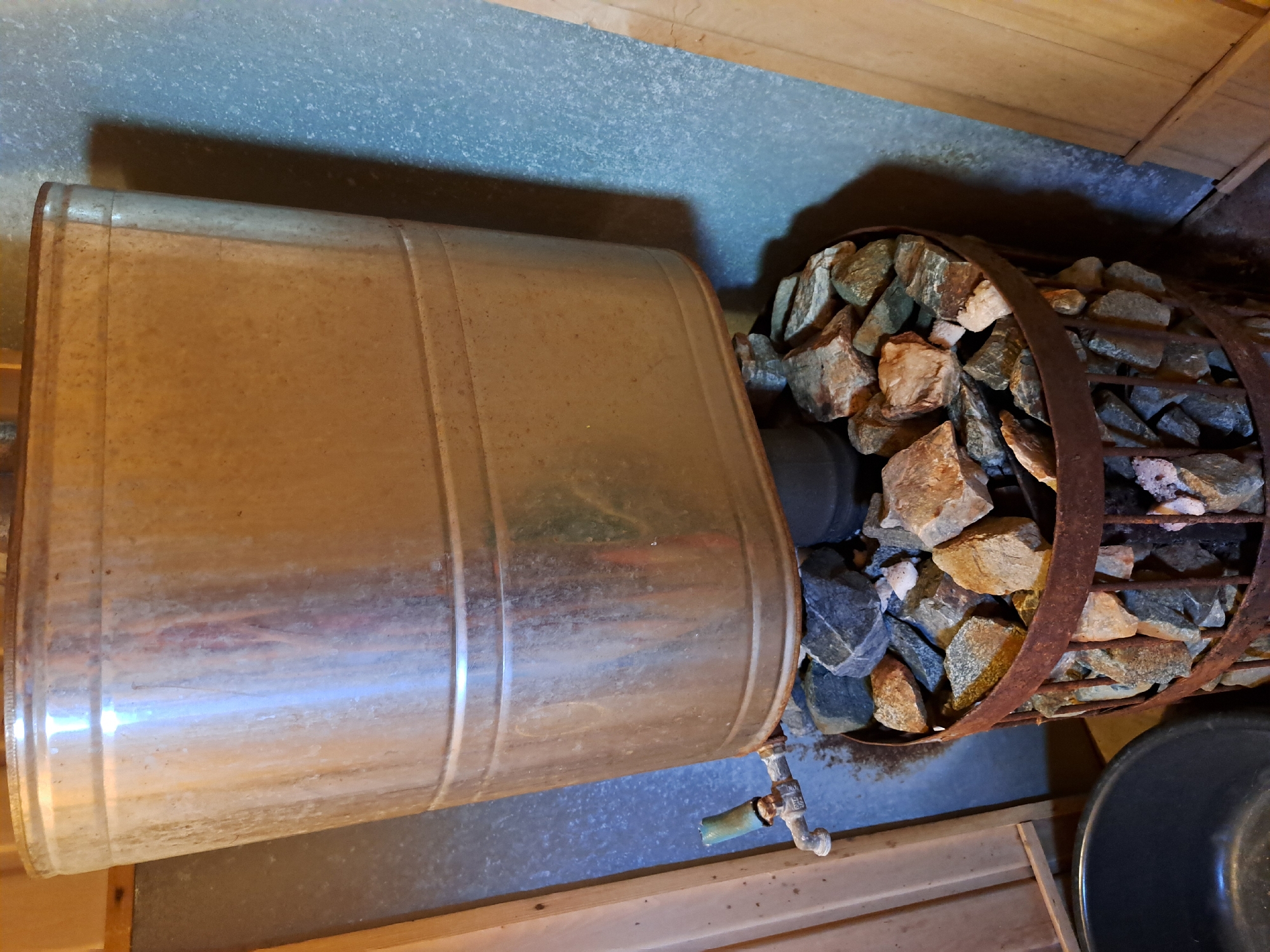 The image shows a close-up, eye-level shot of a rustic sauna heater. The main object is a large, galvanized metal bucket, appearing aged and slightly stained, mounted on a cylindrical structure filled with dark, gray, and tan rocks. The bucket has a spout with a green handle on its lower left side. The rock-filled structure is made of rusty metal bands and vertical supports, creating a cage-like effect. The background consists of a light blue, textured wall on the right and vertical wooden planks on the left. A black plastic basin is partially visible in the lower left corner. The lighting suggests an indoor environment, possibly a sauna or similar rustic setting.
