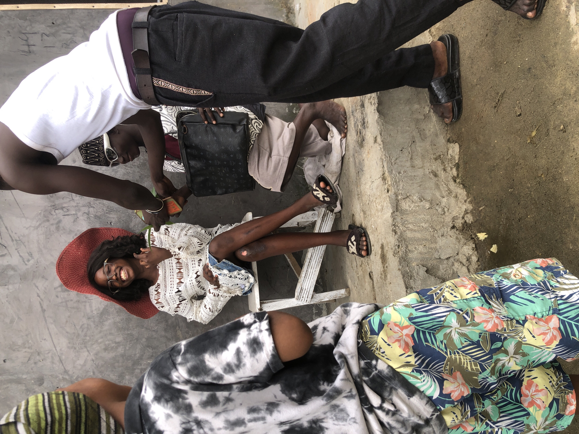 In Ado, Nigeria, a group of four individuals are gathered outdoors near a concrete wall. A young person wearing a tie-dye shirt and floral shorts stands facing away from the camera, their back to the viewer. To their right, a young woman with dark curly hair sits on a white stool, wearing a large red hat and a white crocheted dress. She is smiling and looking towards someone off-camera, holding up a hand with fingers extended. To the right of the seated woman, a person wearing a white tank top and dark pants stands, holding a black bag. Another individual, seen partially, is wearing a patterned shirt and a cap, and is positioned in front of the person in the white tank top. The ground is concrete, wet in places, with some debris scattered around. The overall atmosphere appears casual and informal.