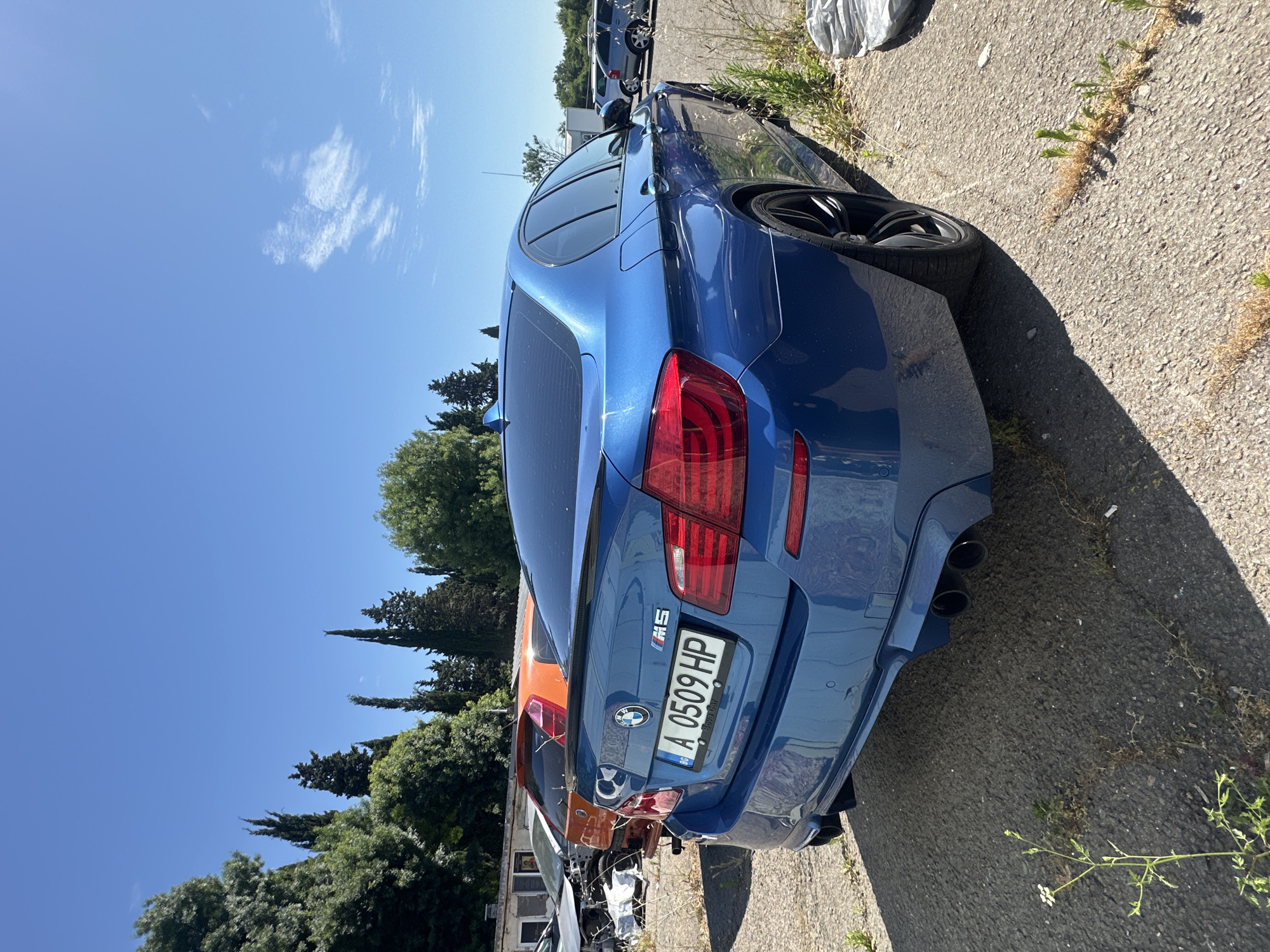 The image depicts a bright, sunny day in an outdoor setting, possibly a junkyard or an abandoned lot, with a clear blue sky overhead. The primary subject is a blue BMW M5 sedan parked on an asphalt surface. The car is viewed from the rear passenger side, highlighting its sleek design, tinted windows, and quad exhaust pipes. A license plate with the characters "BG A 0509 HP" is clearly visible on the rear bumper.
In the background, several other vehicles are visible. To the left of the BMW, an orange hatchback and a damaged car with its front end dismantled are partially obscured. Further in the background, more cars, including a silver hatchback, are parked. The environment is characterized by a mix of paved ground and overgrown vegetation, with several trees lining the perimeter. The overall impression is one of a neglected or storage area for vehicles under a clear, cloudless sky.