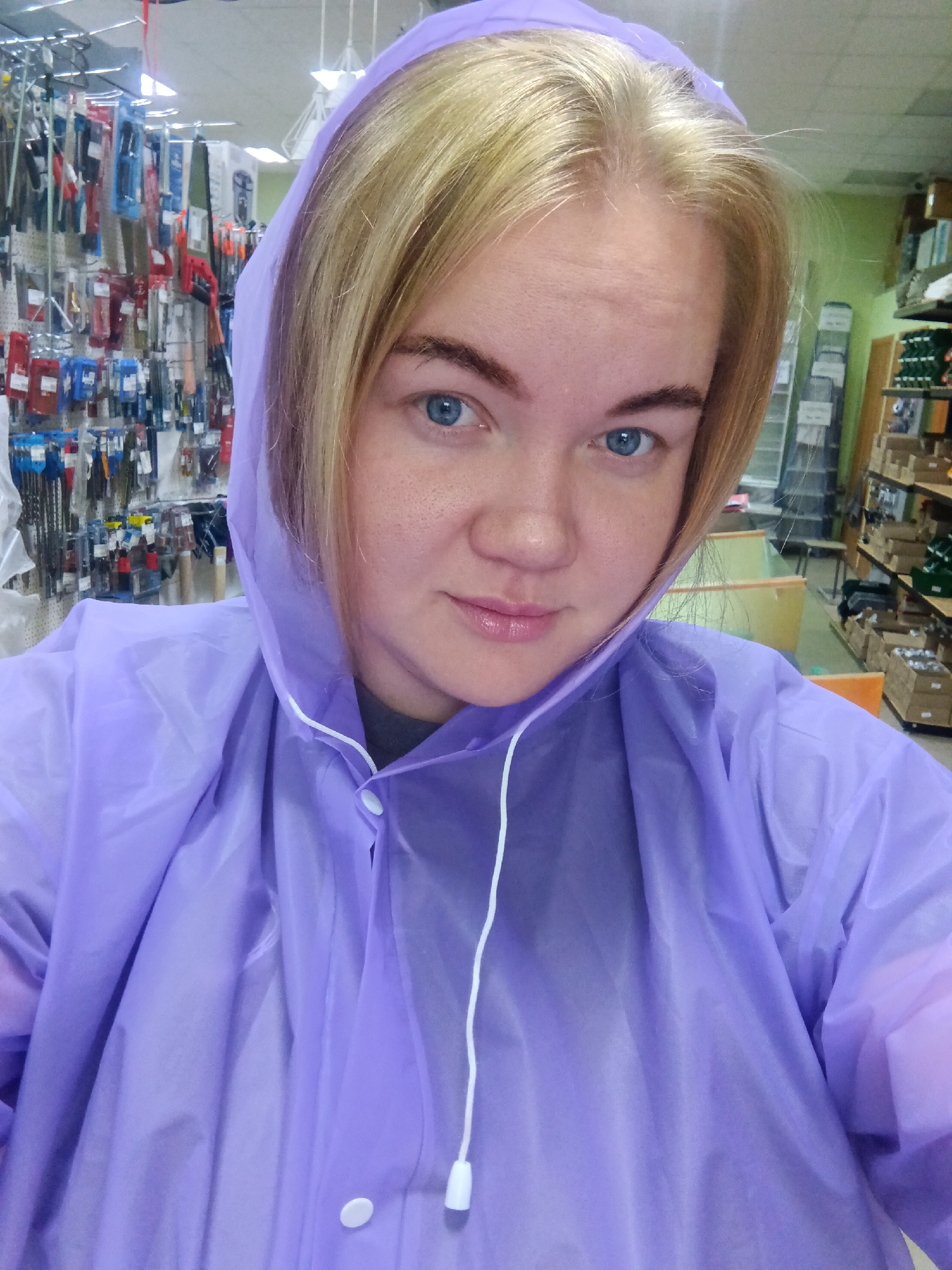 A young woman with blonde hair and blue eyes is taking a selfie in a store. She is wearing a light purple hooded raincoat. The background shows shelves stocked with merchandise, suggesting she is in a retail environment, possibly a hardware or general store. The lighting is bright and artificial. The exact location within the city of Фоки, Russia, is not discernible from the image alone.