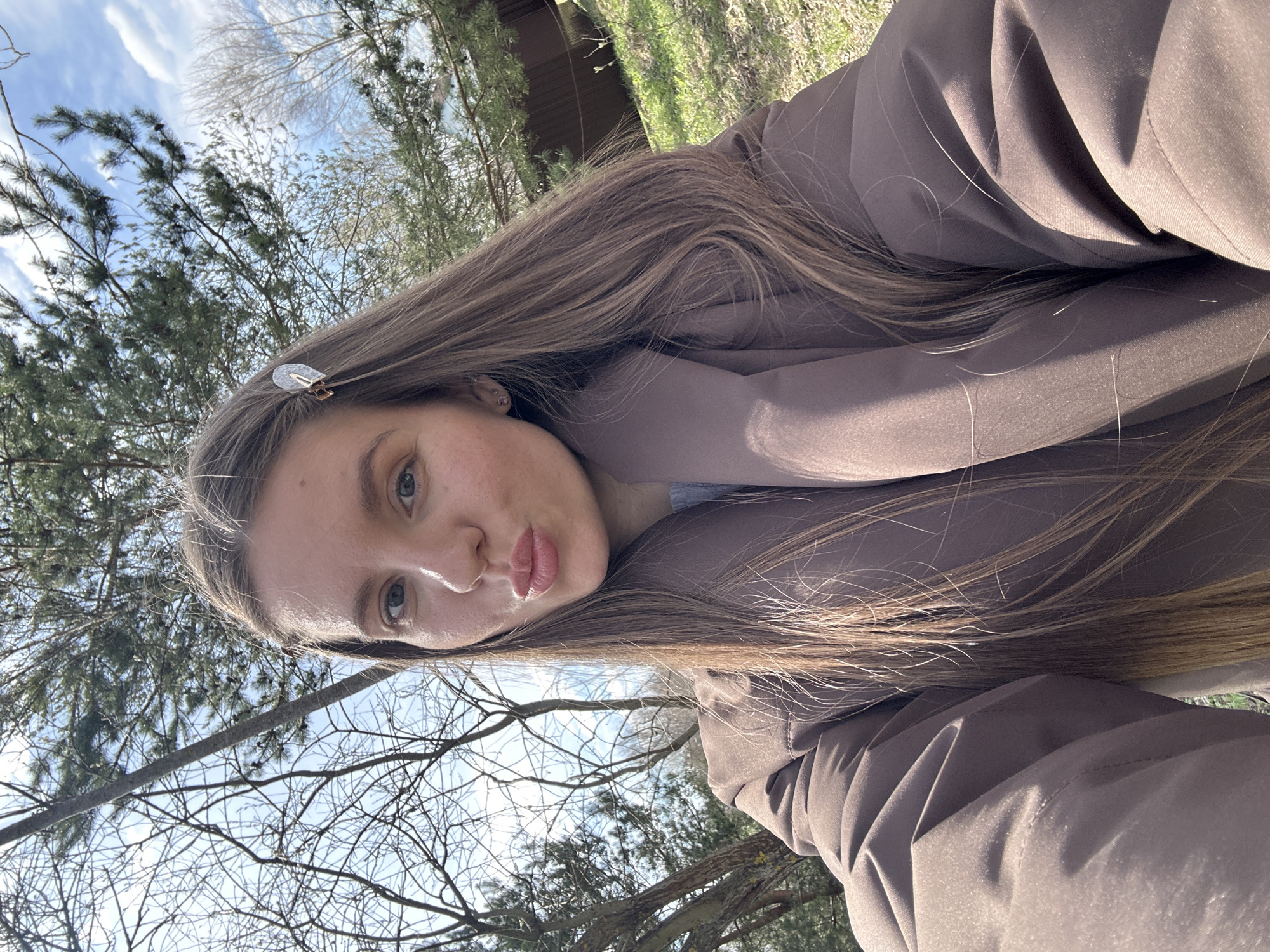 A woman is holding the camera and taking a picture outside. The background shows a building or structure with trees, foliage, and open sky. The location is difficult to assess with accuracy but given the location Мещёры, Russia, it is likely to be a wooded area. The woman is wearing a jacket of neutral color. She has long brown hair with a hair clip. She is looking at the camera and making a kissing face.