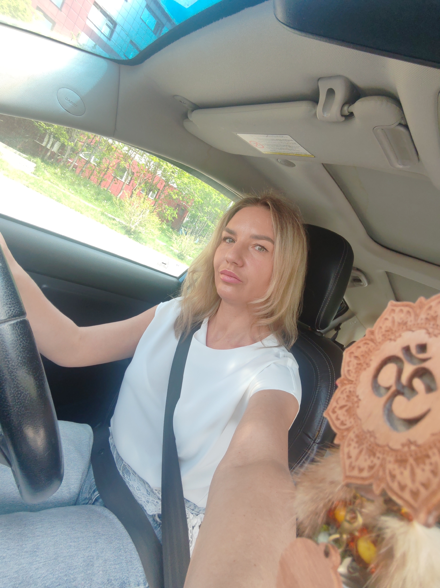 The image shows a woman with blonde hair driving a car. She is wearing a white top and blue jeans. The car's interior is visible, including the steering wheel, dashboard, and a sun visor. The woman is looking at the camera and smiling. A decorative item, possibly a dreamcatcher with an Om symbol, is hanging from the rearview mirror. The background shows a street scene with buildings and trees, suggesting an urban environment. The image appears to be a selfie taken inside the car.
