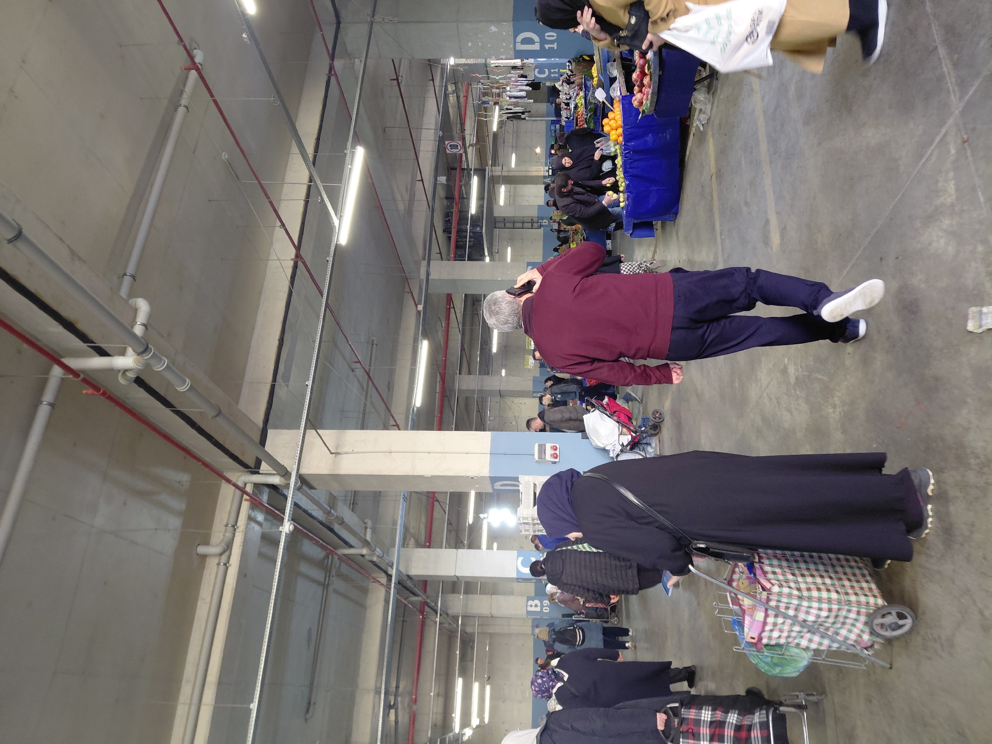 This media file captures an indoor market scene, likely a bazaar, set within a utilitarian concrete structure resembling a repurposed parking garage or basement in Üsküdar, Turkey. The setting features exposed white PVC pipes, red fire suppression pipes, black electrical conduits, and numerous fluorescent strip lights running across the concrete ceiling and walls.
The primary activity is shopping. Numerous individuals are present, mainly adults, navigating between market stalls. In the mid-ground, a man with gray hair, dressed in a maroon sweater and dark pants, is seen walking while speaking on a mobile phone. Ahead of him, a woman in a black abaya and head covering pushes a metal shopping cart laden with a plaid bag and other groceries. Further along the aisle, other shoppers are gathered around various stalls, identifiable by their blue coverings, displaying fresh produce such as oranges and apples. Another person is visible near a stall in the upper right, holding a white shopping bag with partially legible green text. Several shopping carts are scattered throughout the scene.
Large concrete support columns, some painted blue and white, partition the space. Visible text on these columns includes "C D 11 10" and "B 09", likely indicating section or aisle numbers. A circular sign bearing the number "10" is also mounted on a vertical pole. The scene conveys a sense of everyday commerce and community activity under artificial lighting, suggesting that the specific time of day is not a factor for the indoor market.