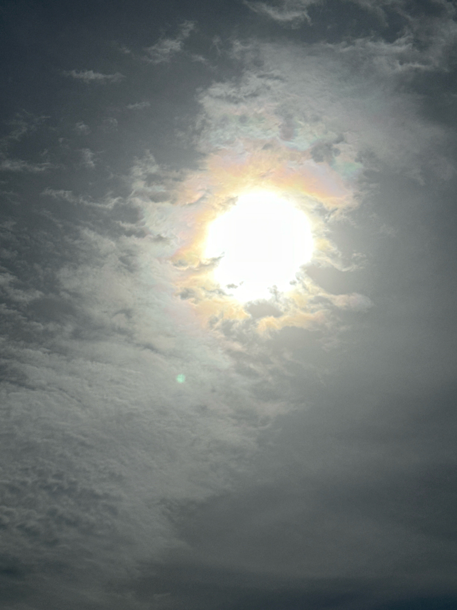 The image displays a sky filled with clouds, dominated by the bright glare of the sun which is partially obscured by them. A faint rainbow effect is visible around the sun. The clouds are varied in texture and density, creating a dynamic and atmospheric scene. The overall color palette is muted, with grays and whites prevailing, punctuated by the intensity of the sunlight. The image quality suggests it was taken outdoors, likely during daytime. There are no discernible landmarks or elements that would definitively place it in Нарацкі сельскі Савет, Belarus.