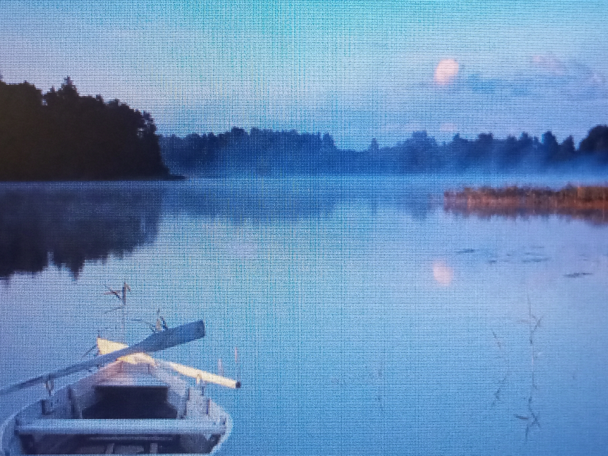 This media file depicts a tranquil lake scene at dawn, likely in Riga, Latvia. The water is calm, reflecting the soft blue and cyan hues of the sky. A dark silhouette of a tree line borders the distant shore, partially obscured by a gentle mist that rises from the water, adding an ethereal quality to the atmosphere.
In the foreground, a small rowboat rests on the water, its weathered grey hull suggesting it has seen many peaceful mornings. Two wooden oars lie within the boat, one resting across the gunwales, ready for a silent journey. The scene evokes a sense of peace and solitude, characteristic of early morning by a secluded body of water.
The time of day is clearly dawn, indicated by the soft, diffused light and the pale sky transitioning from deep blue to lighter shades, with a faint hint of a pinkish cloud or celestial body visible in the upper right quadrant. The weather appears calm and cool, with the mist suggesting humidity and a lack of wind. There are no visible people or text within the image.