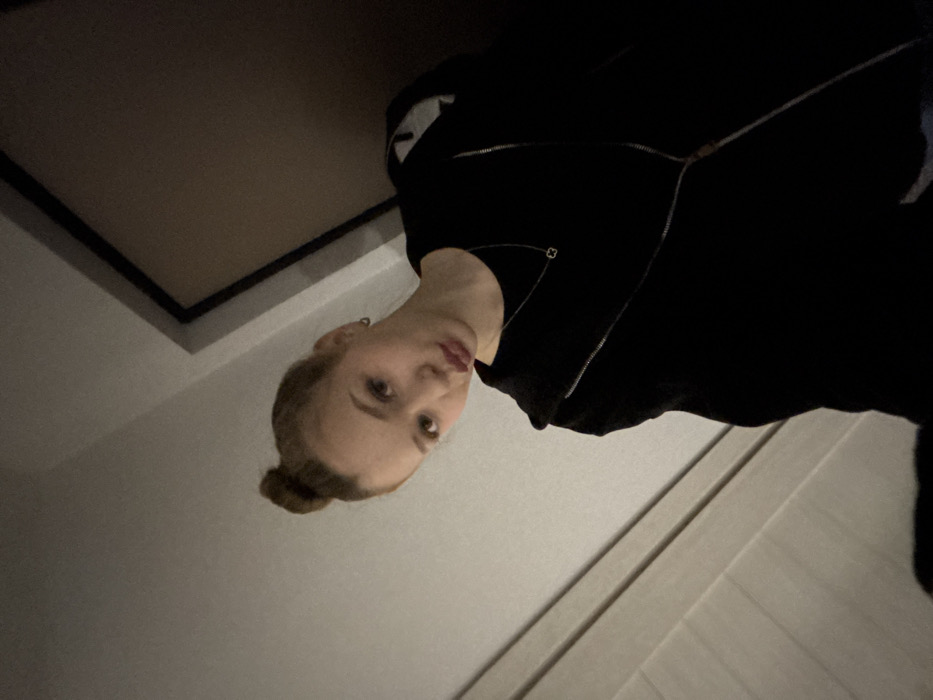 A young woman with fair skin and blonde hair in a bun is depicted indoors. She is facing the camera, wearing a black zip-up jacket and a small hoop earring. Behind her, a white paneled wall stretches diagonally. The top left corner features a dark-edged ceiling panel. The overall lighting is soft, creating subtle shadows on the walls. The image is taken from a slightly elevated angle, giving the viewer a unique perspective.