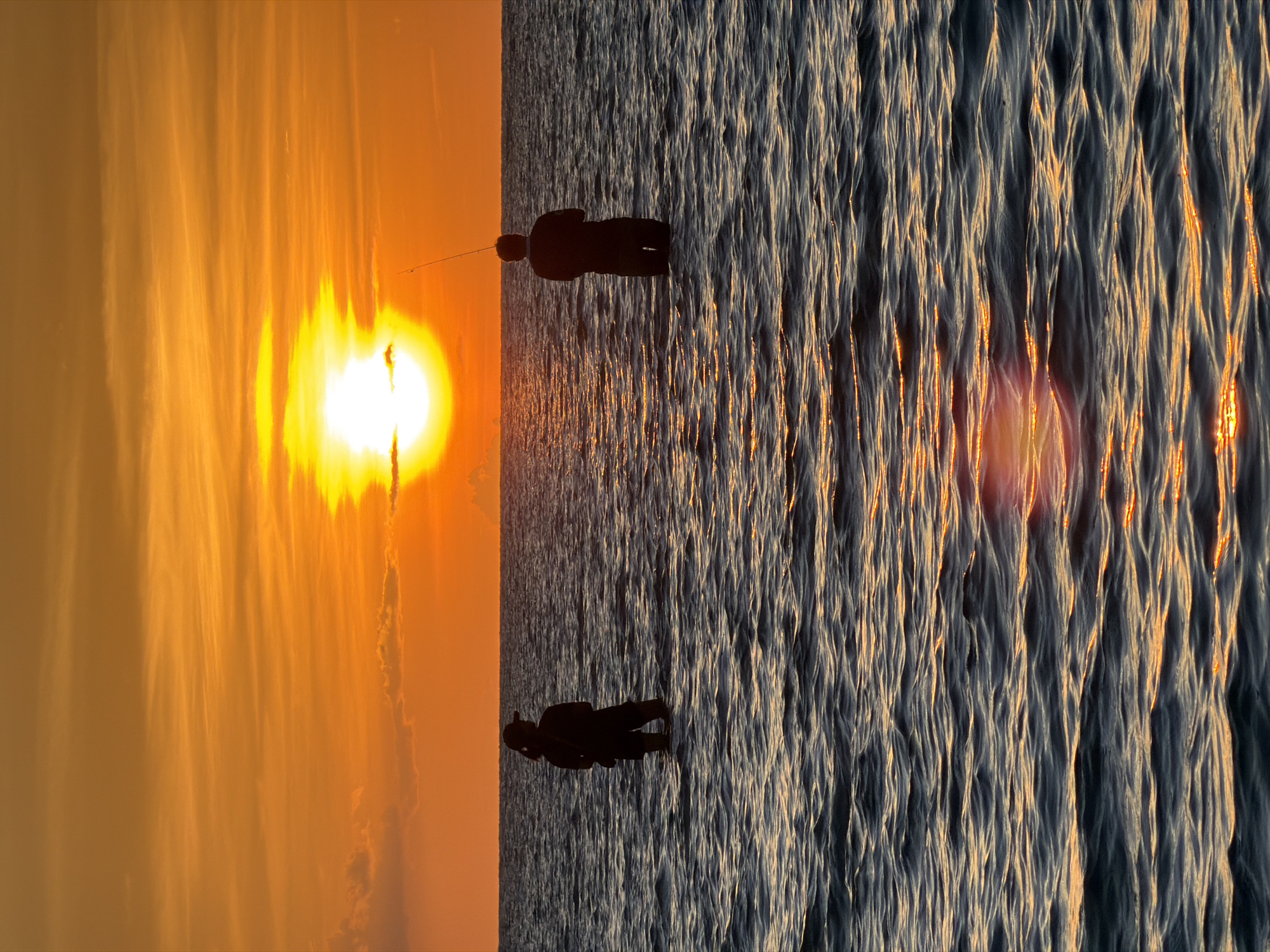 Two individuals are standing in the ocean near Baan Plai Laem, Thailand, engaged in fishing during sunset. The sun, a bright orb, is positioned near the horizon, casting a warm, orange glow across the sky and reflecting on the water's surface. The sea exhibits gentle waves, with ripples catching the sunlight. One person, on the left, is wearing a hat and appears to be looking out towards the sea. The other person, on the right, holds a fishing rod, actively fishing. The overall scene depicts a peaceful and serene moment of recreational activity against a dramatic natural backdrop.