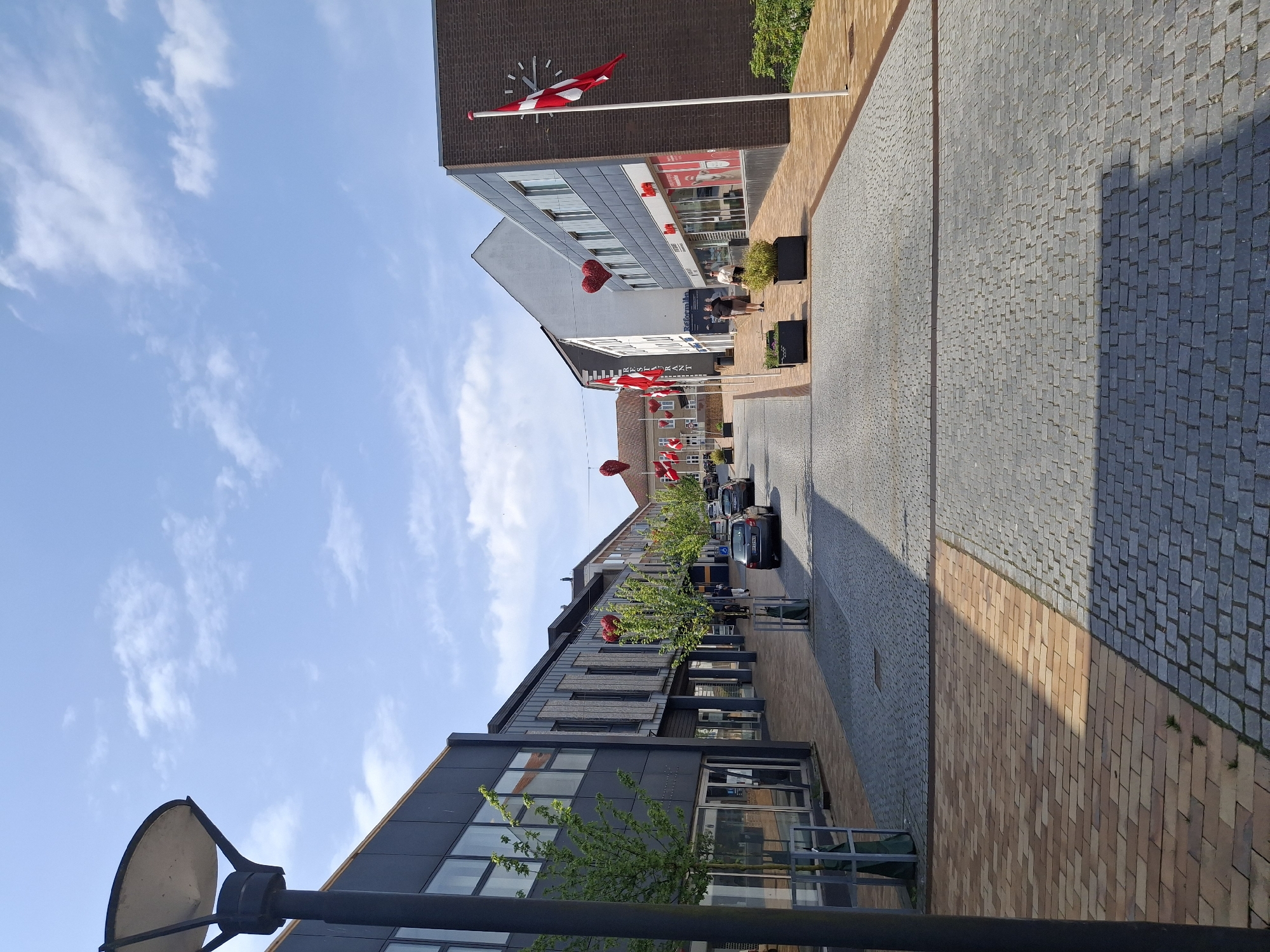 The image depicts a street scene in Aabenraa, Denmark, viewed from a low angle. The street is lined with buildings of varying heights and architectural styles, including modern structures with glass windows and older brick buildings. Danish flags are prominently displayed on buildings and strung across the street. Vehicles are parked along the street. People are visible walking near a building with the inscription "SYDBANK". The sky is blue with scattered clouds. The street surface is paved with cobblestones and bricks. A lamppost is partially visible in the foreground.