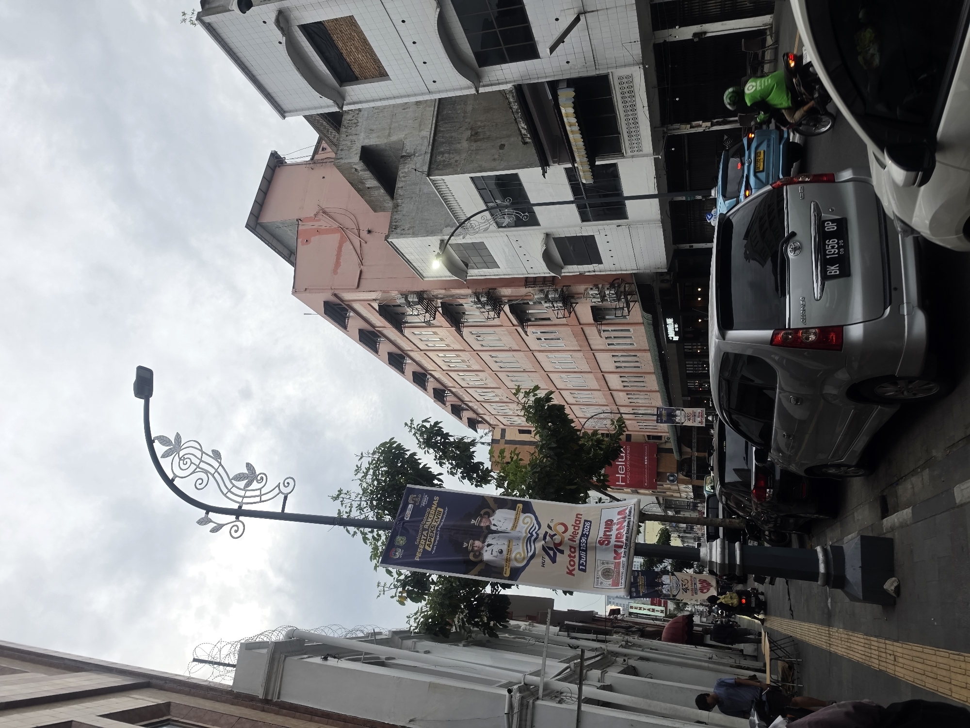 The image depicts a street scene in the city of Medan, Indonesia. A light gray sedan is parked on the right side of the street, facing away from the camera. Behind it, a blue car is driving away, and a motorcyclist in a green jacket is also moving away. The street is lined with buildings of various architectural styles and colors, including a pink, multi-story building and white buildings with tiled facades. A lamppost with decorative metalwork stands prominently in the foreground, displaying a banner. The banner appears to be a celebration of "HUT 435 KOTA MEDAN" which translates to "435th Anniversary of the City of Medan." There are also some pedestrians visible on the sidewalk on the left side of the street, and more parked cars. The sky is overcast with clouds. The scene suggests a typical urban environment with commercial and residential buildings, traffic, and public decorations.