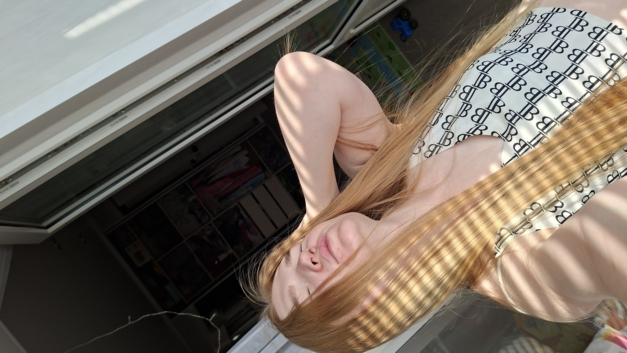 Here's a detailed description of the image:
**People:** A fair-skinned person with long, straight, reddish-blonde hair is the primary subject. The individual is facing towards the viewer, with their head resting on their arm.
**Subjects:** The person is wearing a top with a pattern of "B" symbols.
**Objects:** The person is near a window, casting horizontal shadow lines across their face and hair. A partially open window with a white frame is visible, revealing the exterior.
**Actions/Interactions:** The person appears to be resting or posing for the picture.
**Environment:** The setting is indoors, near a window providing natural light. Outside the window, there is a shelf containing several objects. The location is most likely in a residential area.
