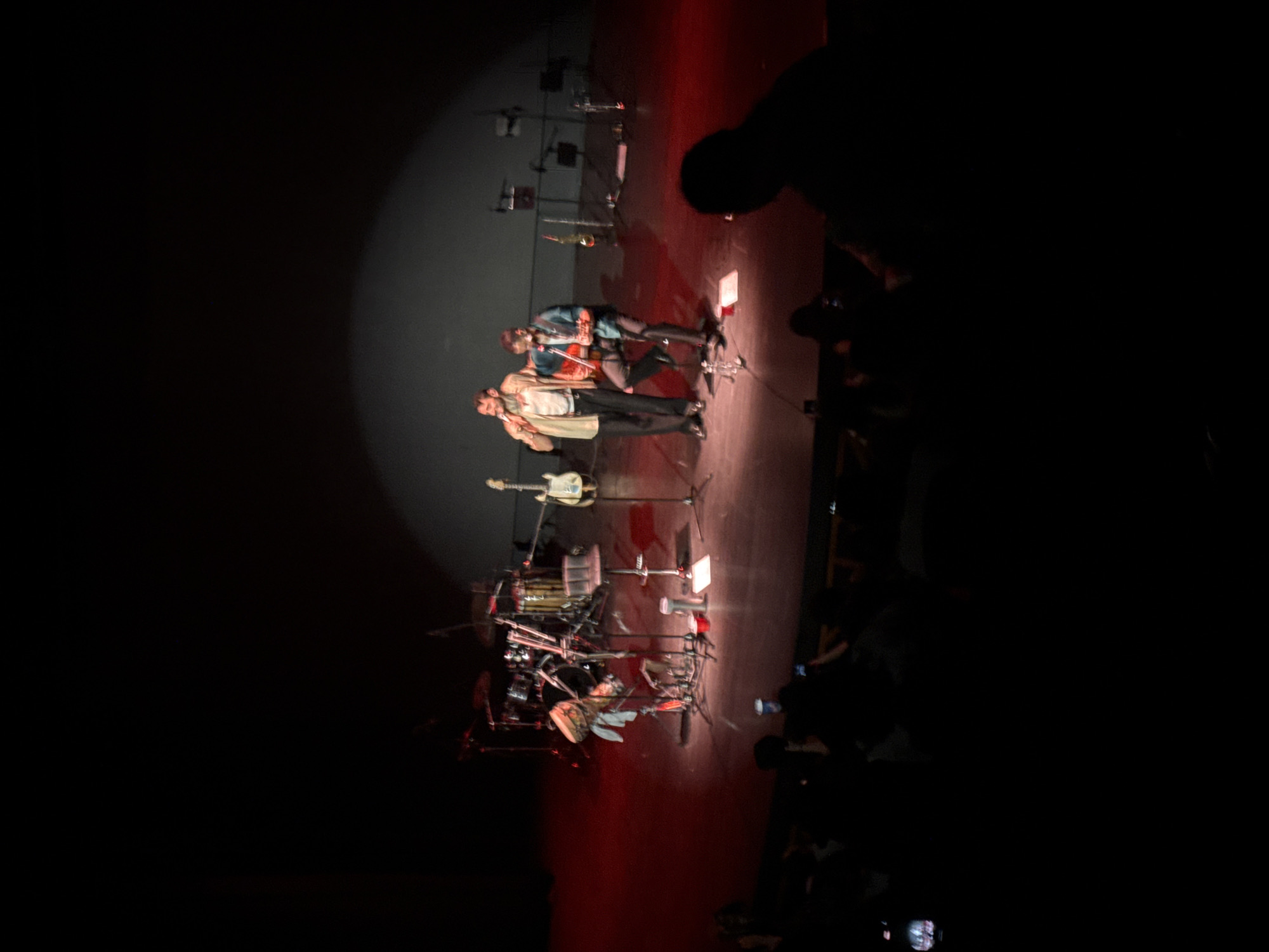 The image depicts a stage scene, likely from a performance or concert, in Tlalnepantla, Mexico. Two individuals are standing center stage under a spotlight. A band's musical instruments, including a drum set and a guitar on a stand, are visible behind them. A blurred audience is visible in the foreground, with some individuals holding up phones. The lighting is dim, with a focused spotlight on the performers and the stage floor illuminated with red light.